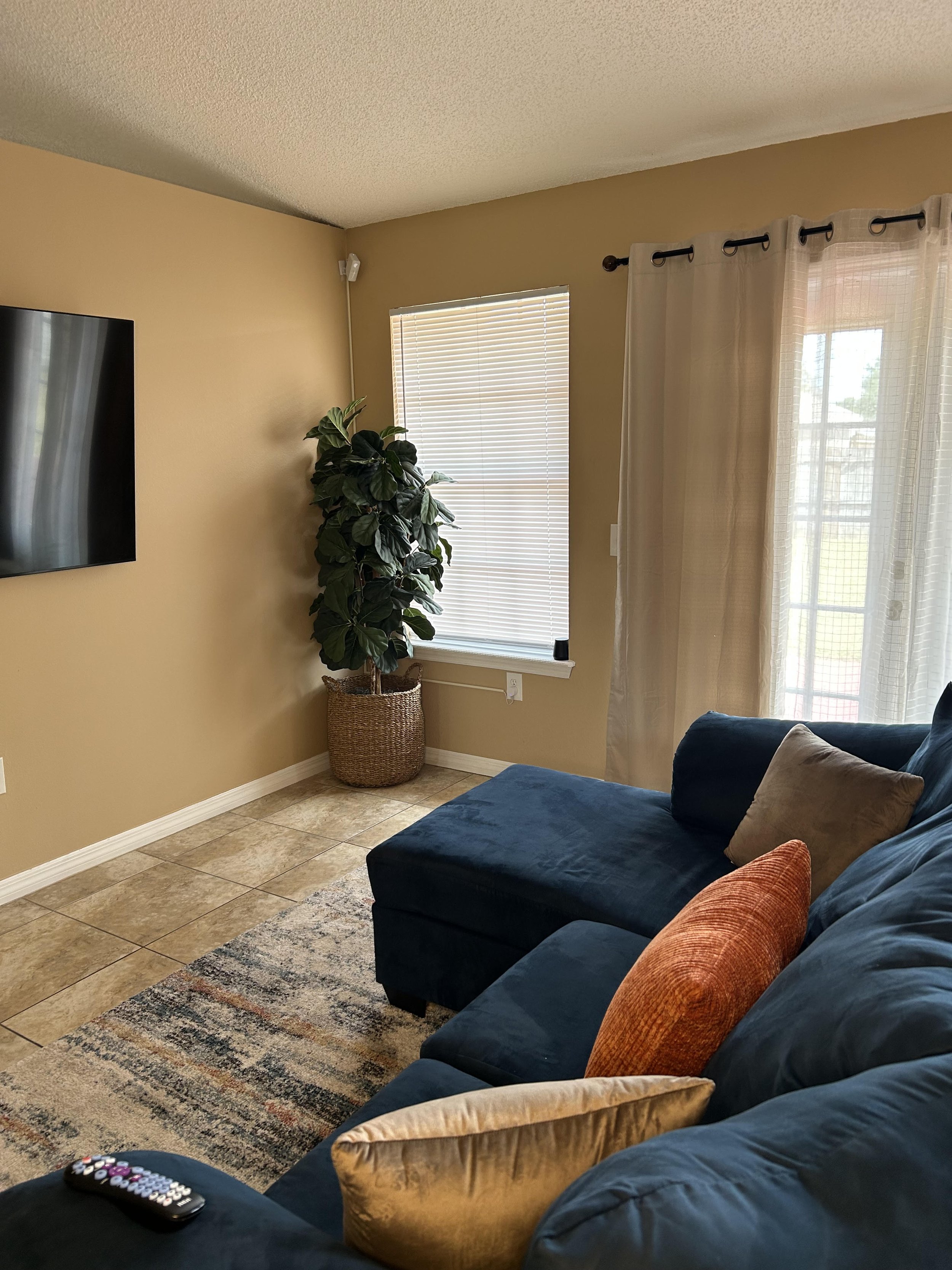 Living room corner with a blue sectional, colorful pillows, potted plant, window, and wall-mounted TV.