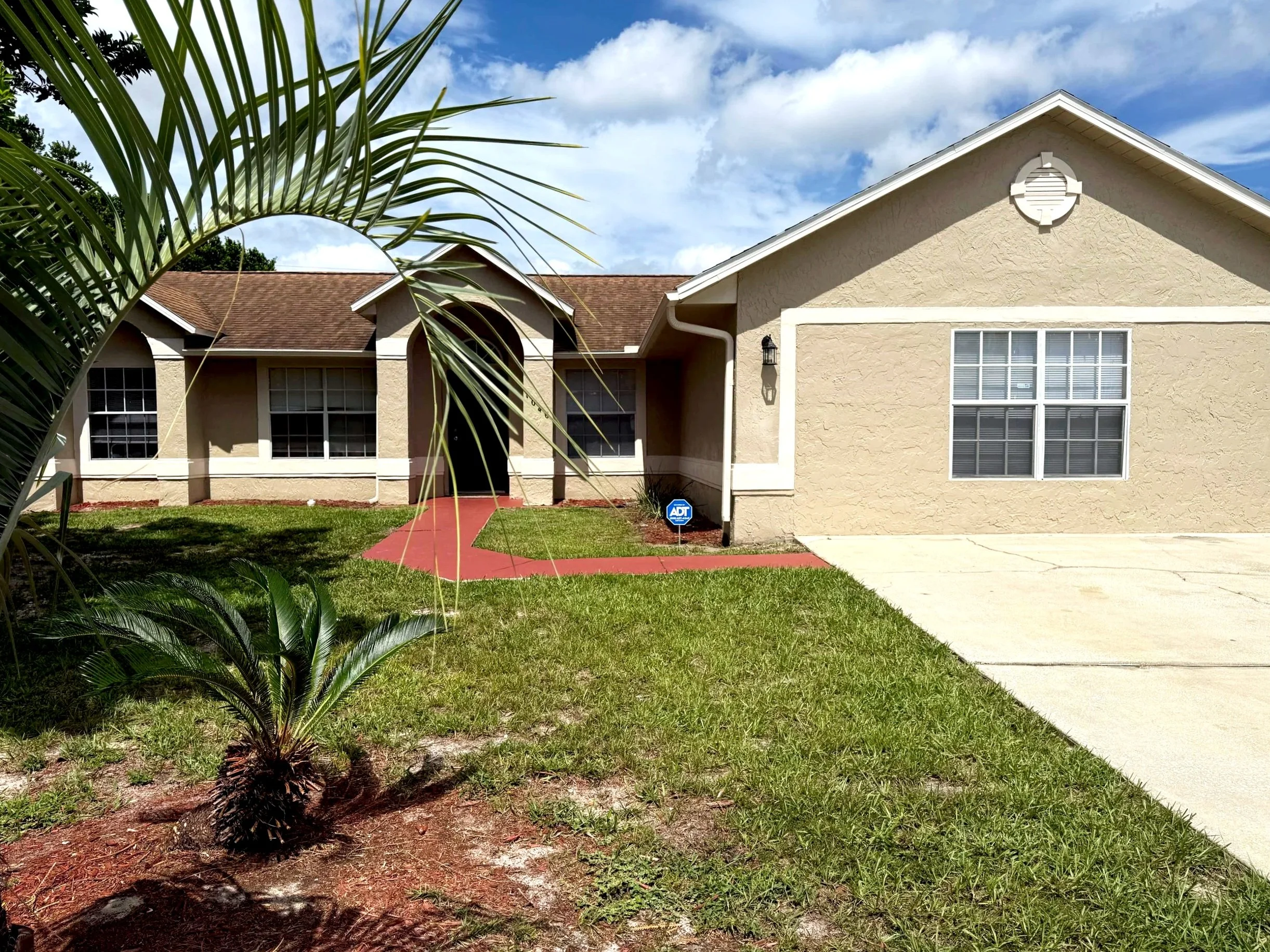 Front of a beige house with brown roof, driveway, walkway, and small landscaped yard.