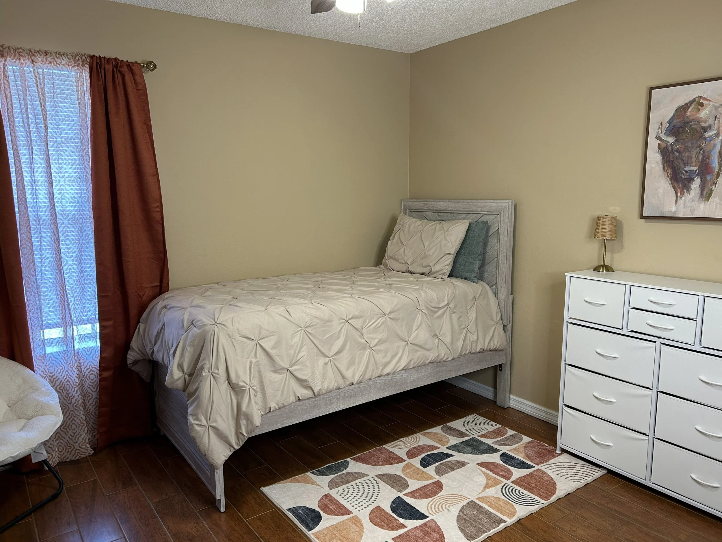 Bedroom with a single bed, white dresser, colorful rug, and a window with red curtains.
