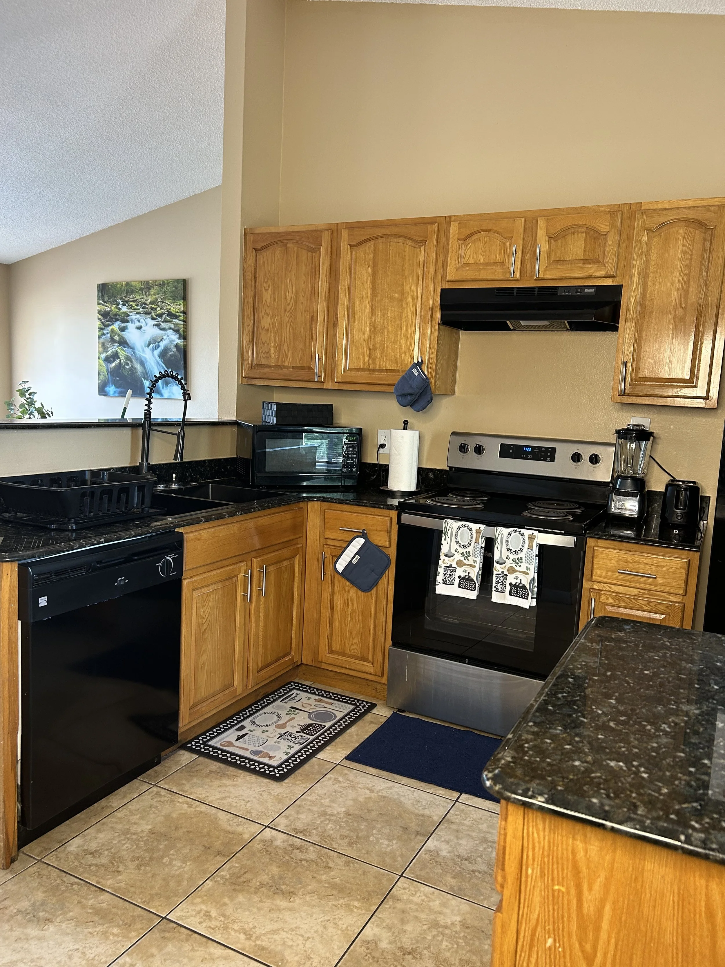 Kitchen with wooden cabinets, black appliances, granite countertops, and a tiled floor with rugs.