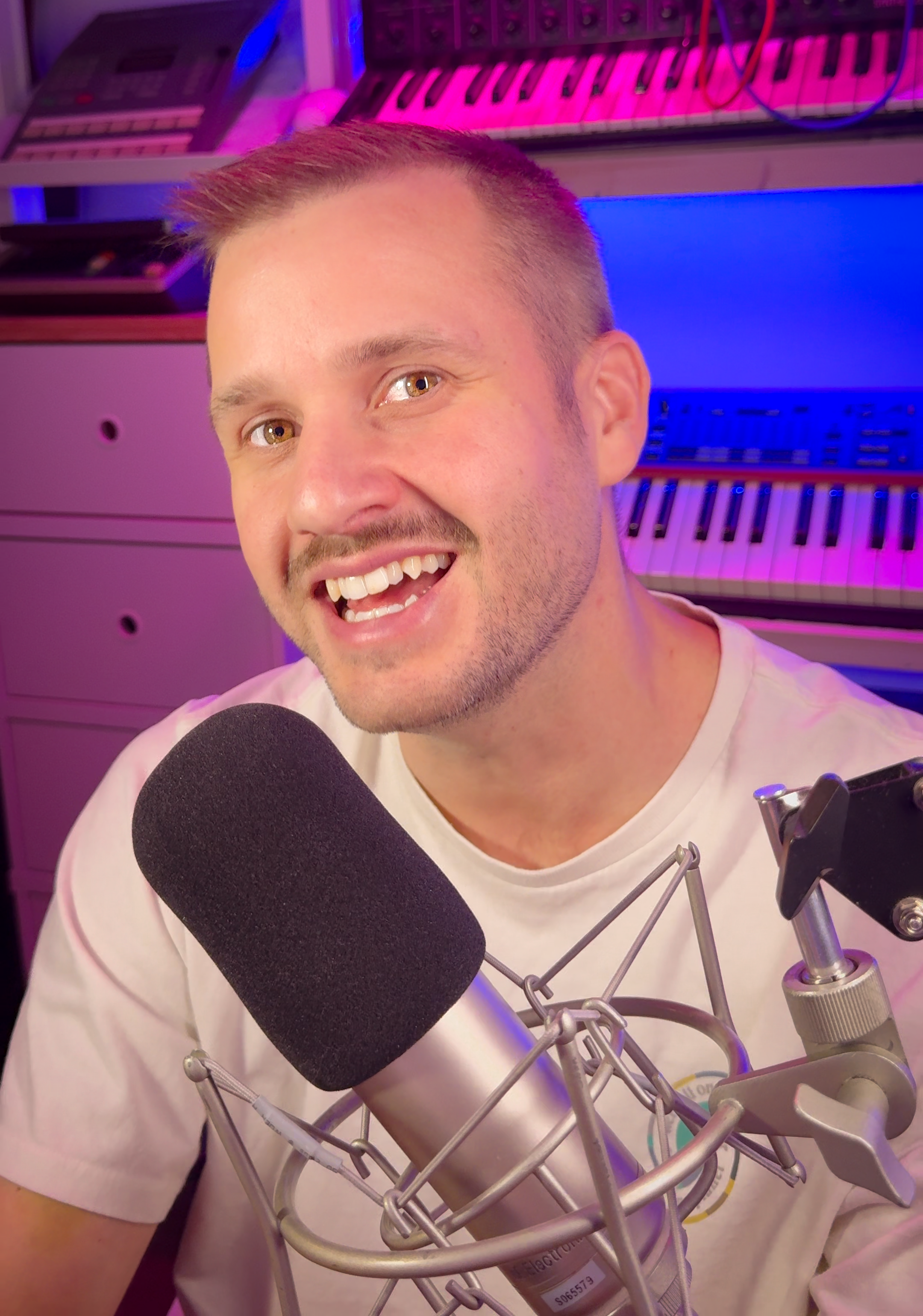 Man met kort haar en een glimlach, voor een microfoon, met muziekinstrumenten op de achtergrond, in een kleurrijke studio