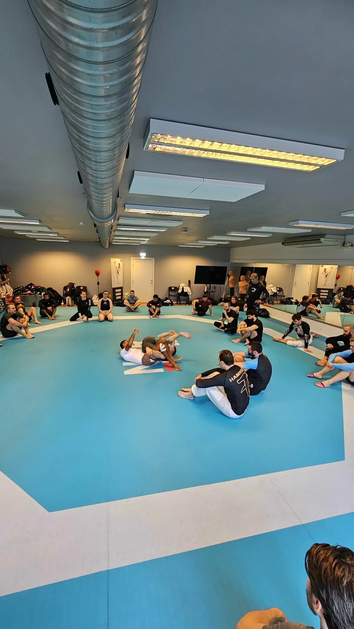 Mennesker trener brasiliansk jiu-jitsu på en innendørs matte med speilvegger.