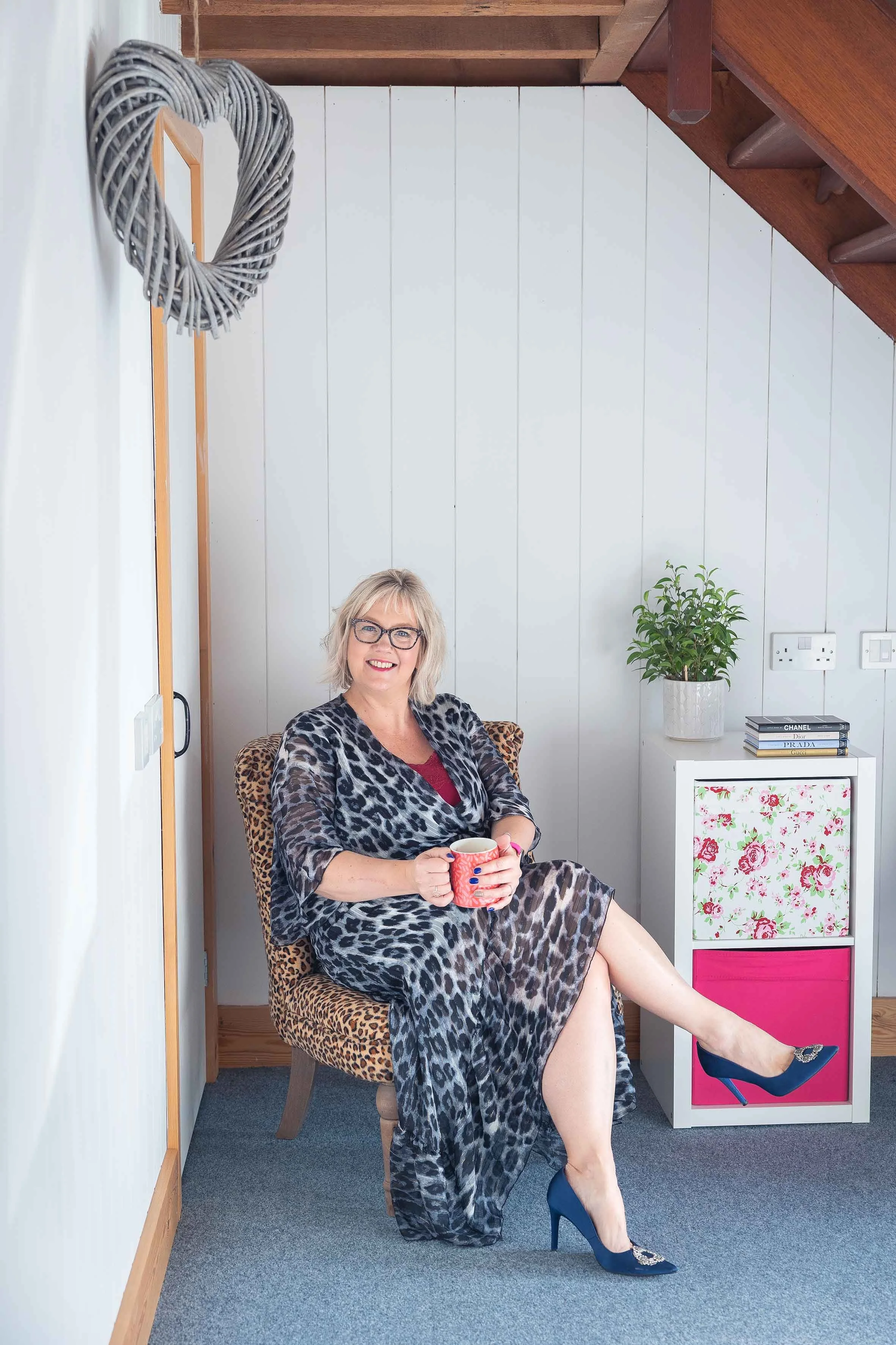 A woman sitting on a leopard print chair in a cozy room, holding a mug, smiling, wearing a leopard print robe, blue high heels, glasses, and a red top underneath. There is a white cabinet with floral design and pink drawer, a potted plant, and a stack of designer books in the background.