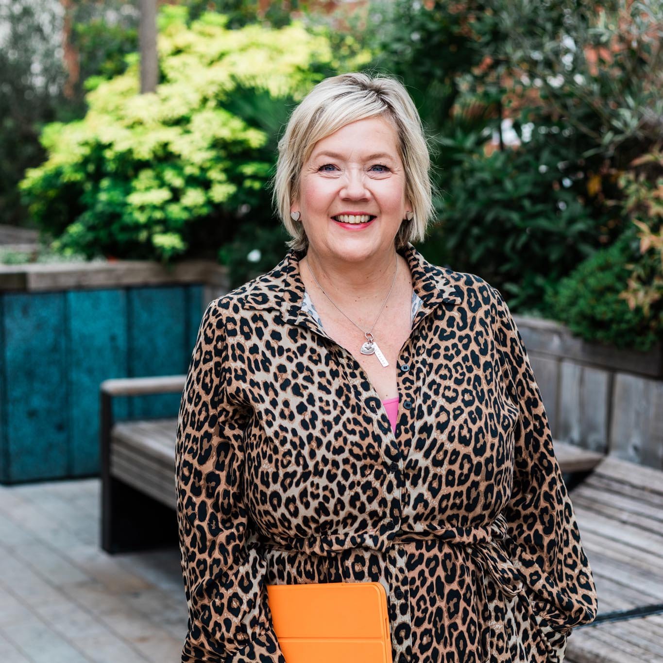 Niki, a smiling woman with short blonde hair, wearing a leopard print dress and jewelry, standing outdoors on a wooden deck with green foliage in the background.