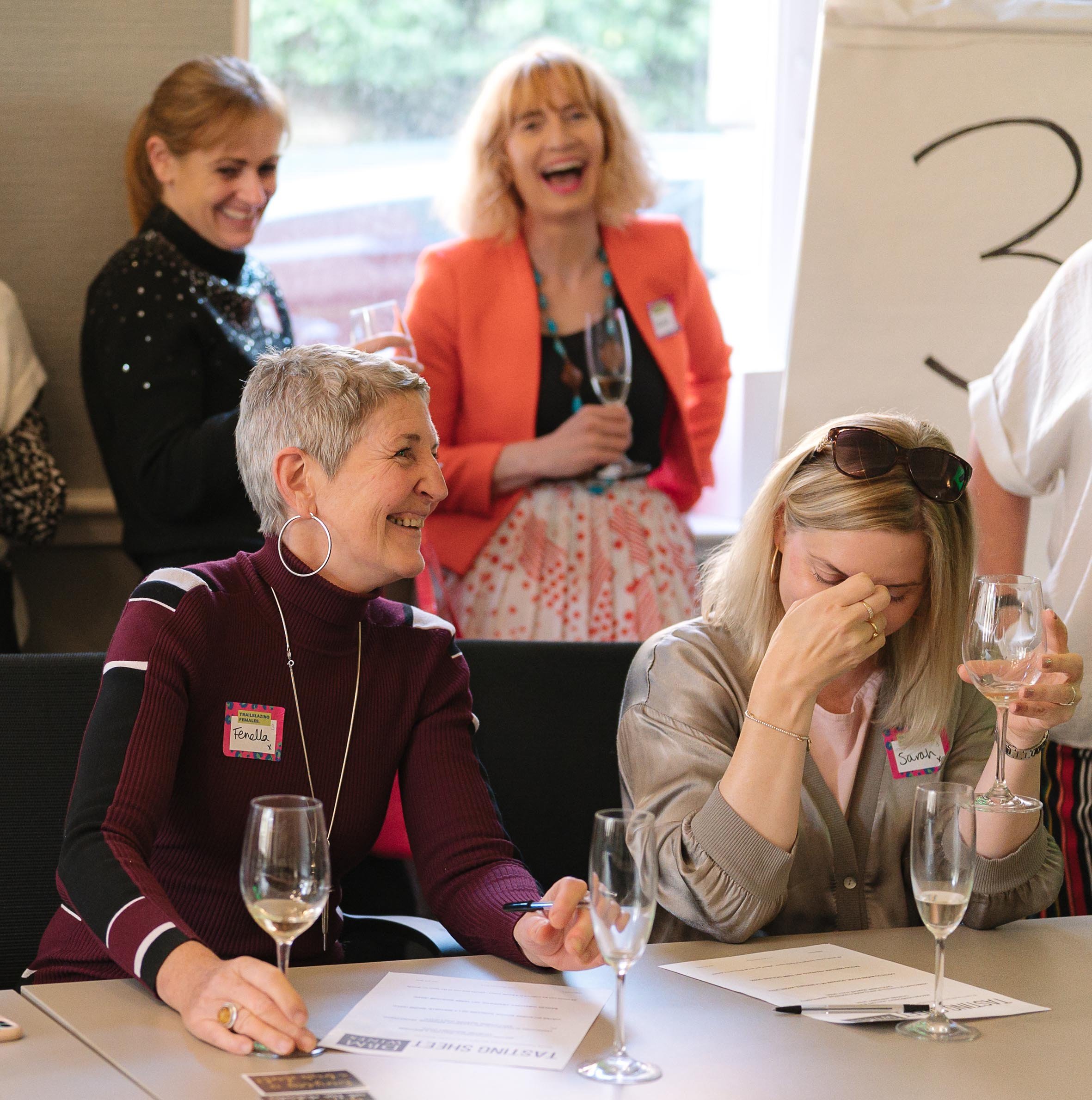 Two women seated at a table, signing papers with glasses of champagne in front of them, at a social or professional event.