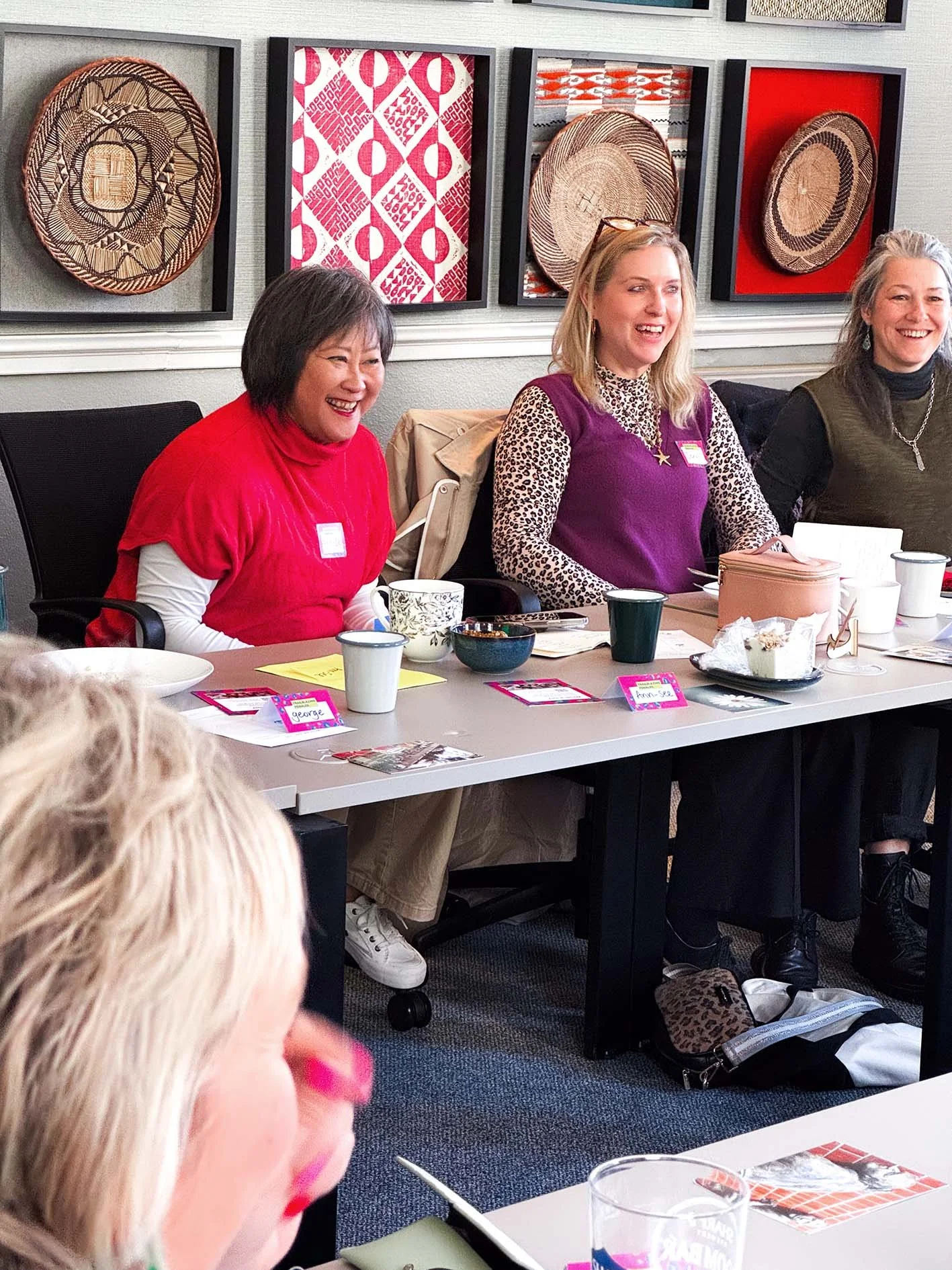 Group of women sitting at a table in a meeting room with artwork on the wall behind them, smiling and engaging in conversation.