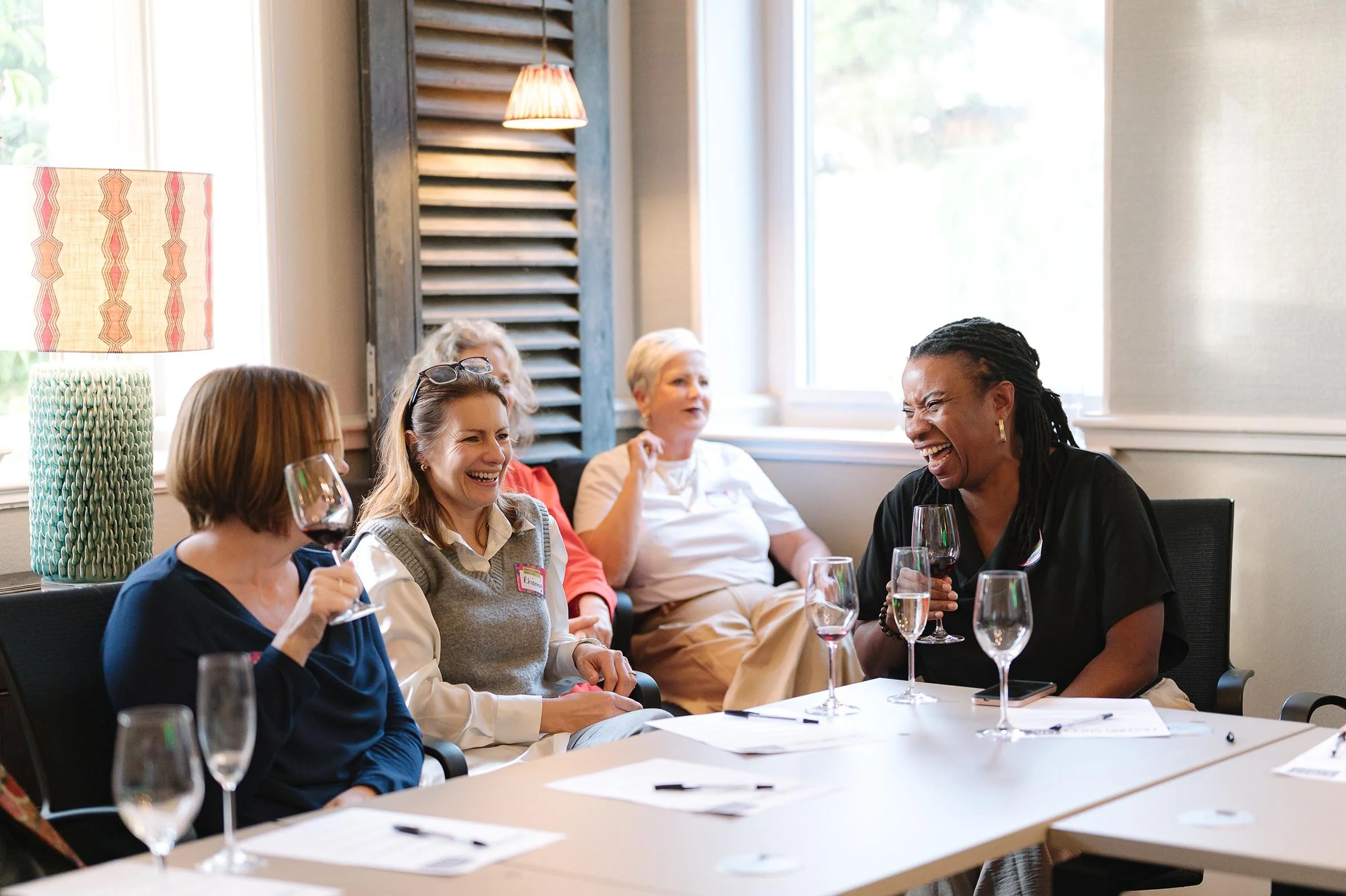 Group of women sitting at a table, laughing and enjoying drinks during a meeting or social event in a well-lit room with large windows and a decorative lamp.