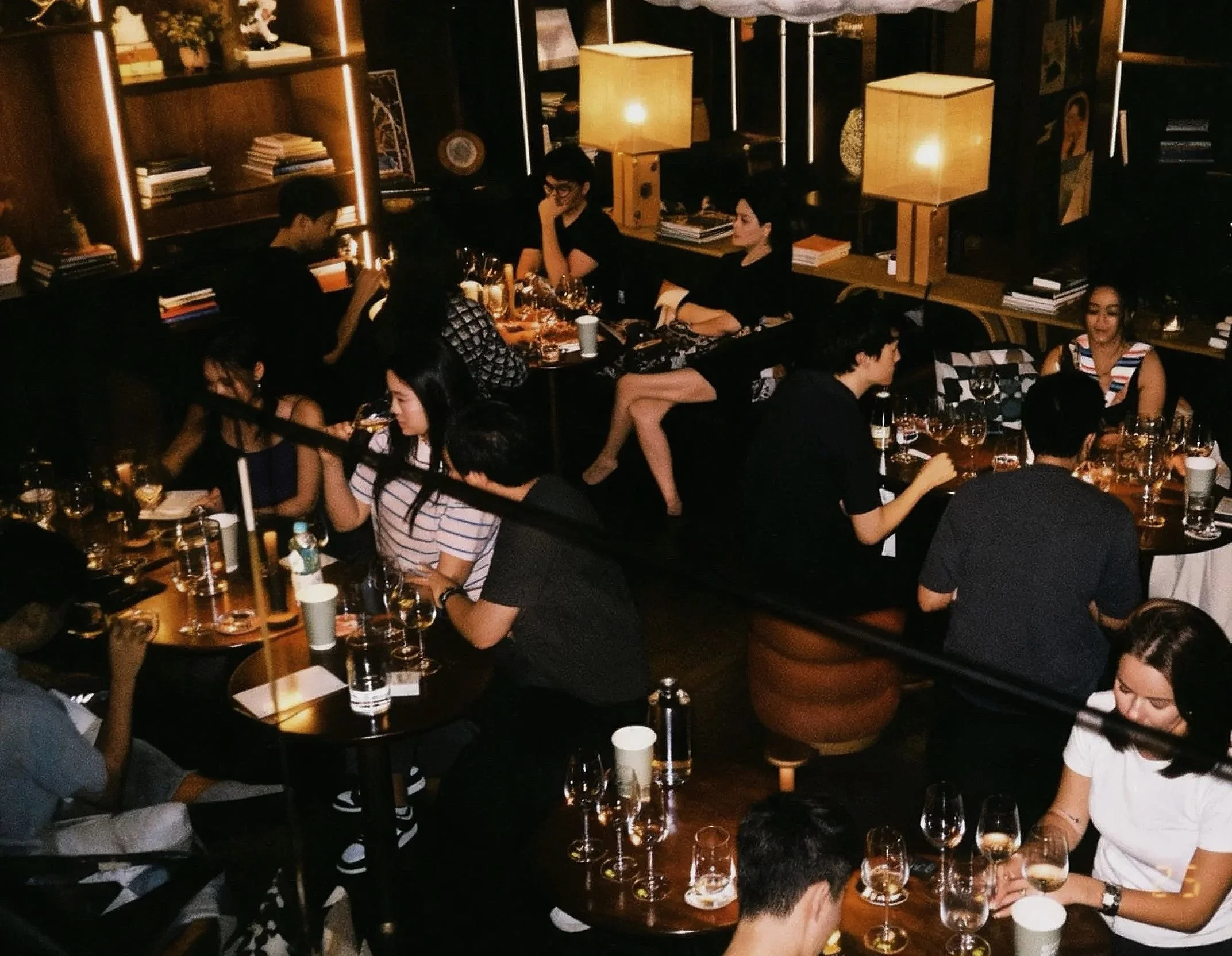 Group of people dining and socializing in a dimly lit restaurant or bar, with glasses of wine on the tables and warm lighting.