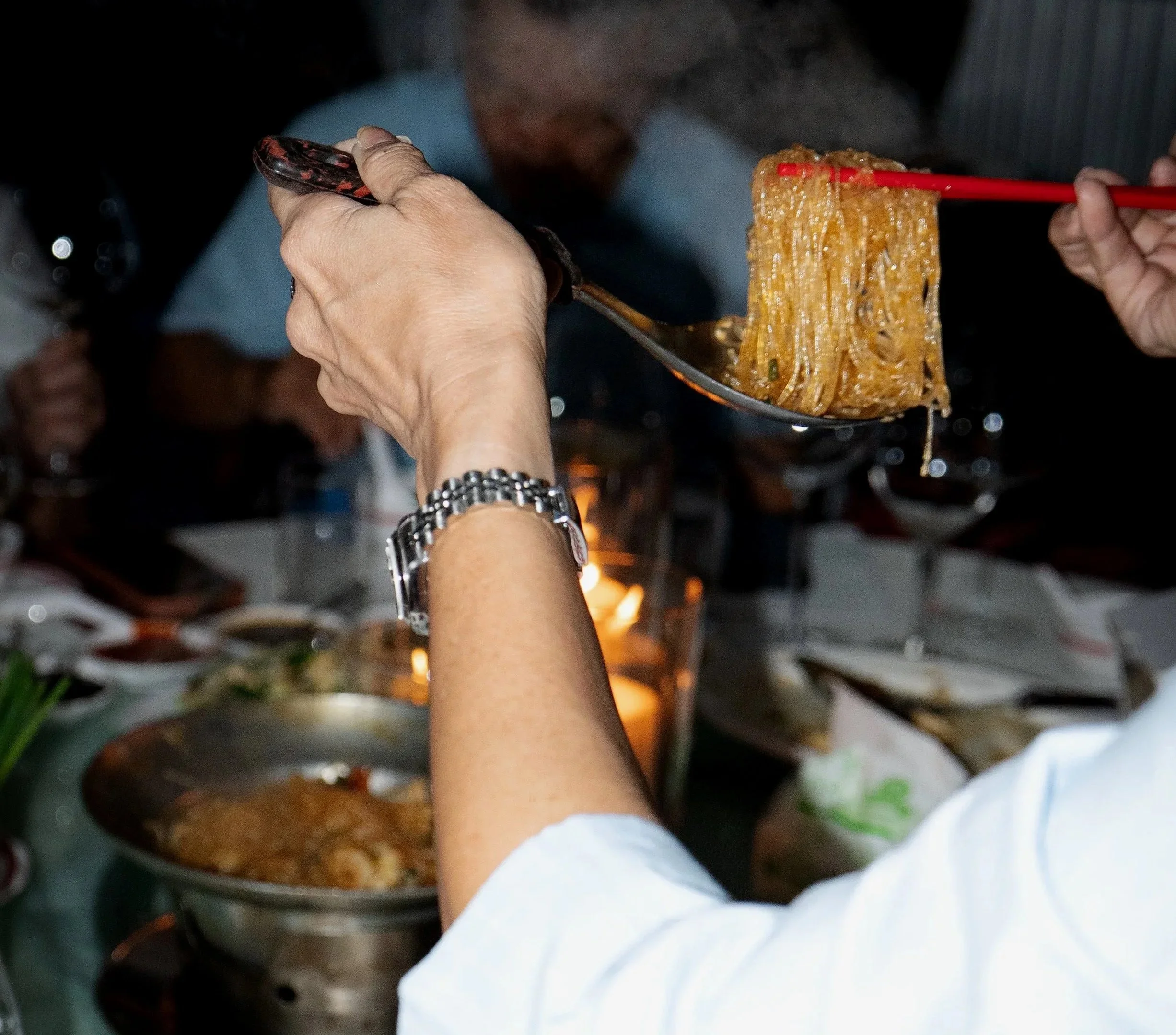 Person holding a spoon with noodles and using red chopsticks to serve noodles at a dinner gathering.