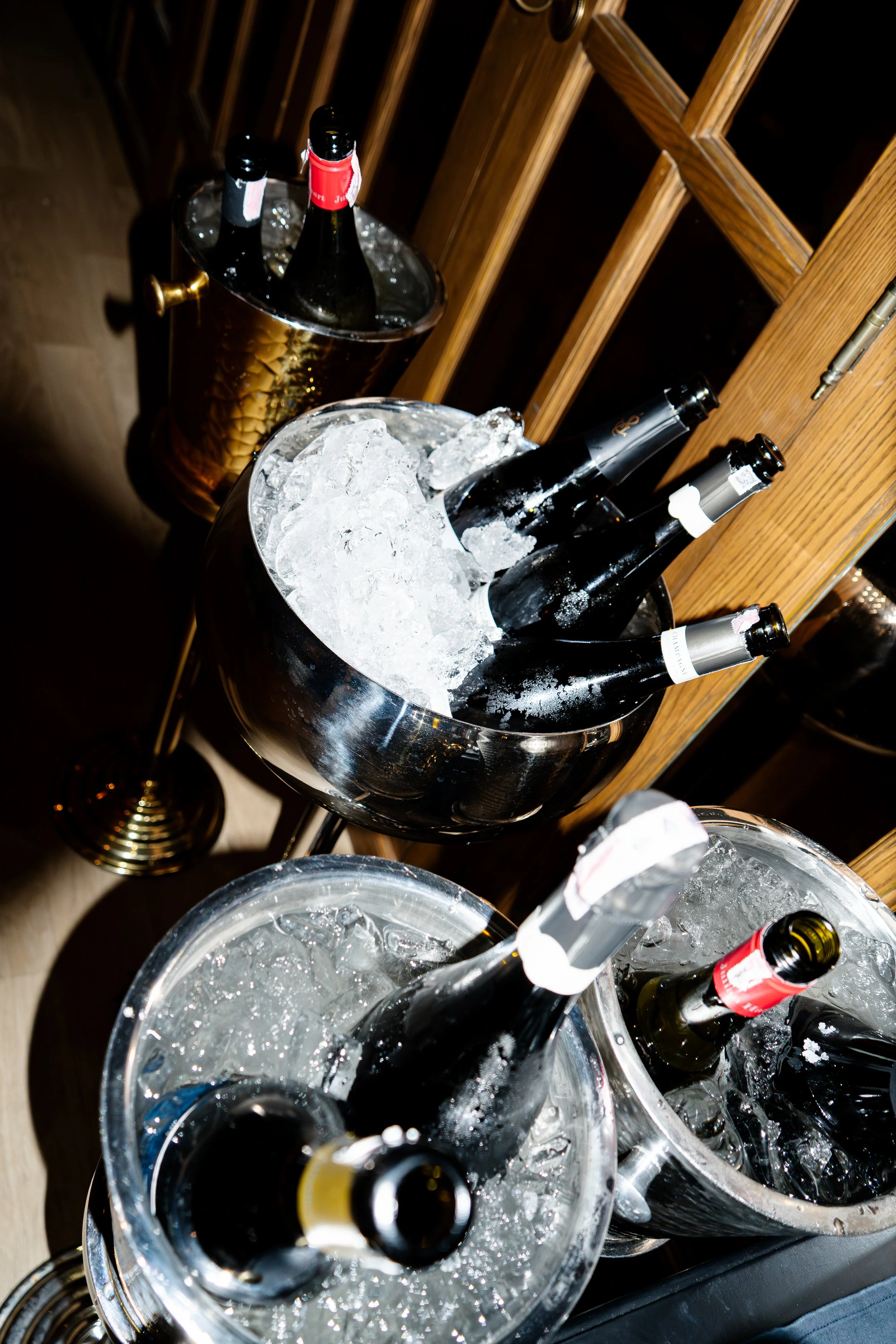 Several bottles of champagne chilling in ice buckets and wine bottles in a bucket of ice, on a wooden bar or counter.