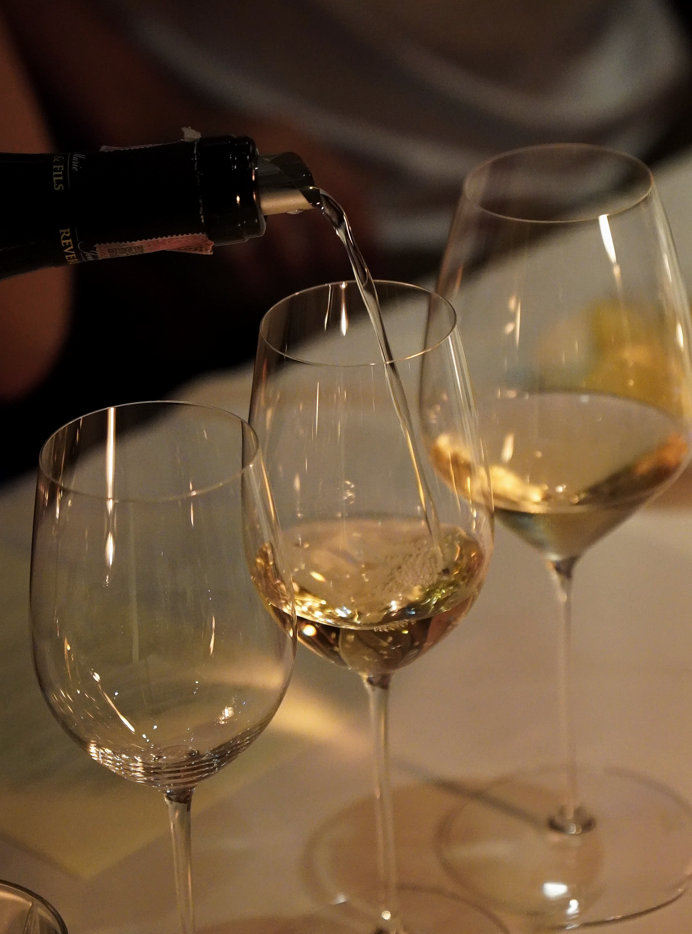 A hand pouring white wine into a glass, with two other glasses of white wine nearby, on a table.