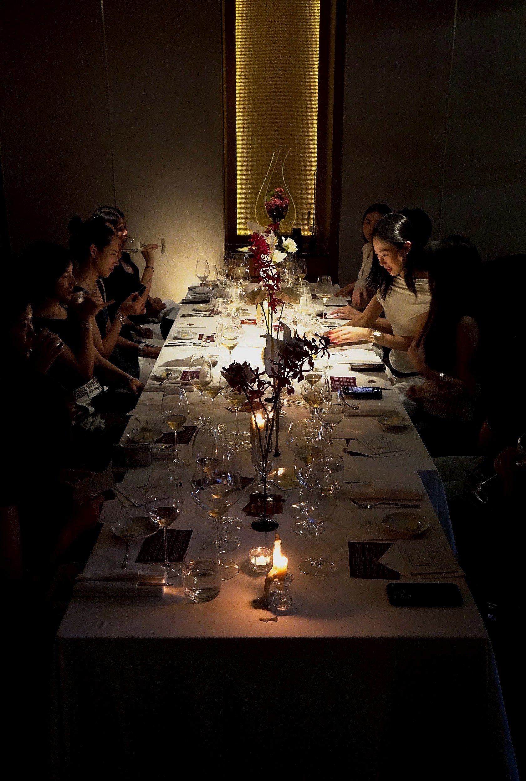 A long dining table set for a dinner party with wine glasses, floral centerpiece, and candlelight, with several women sitting around it in a dimly lit room.