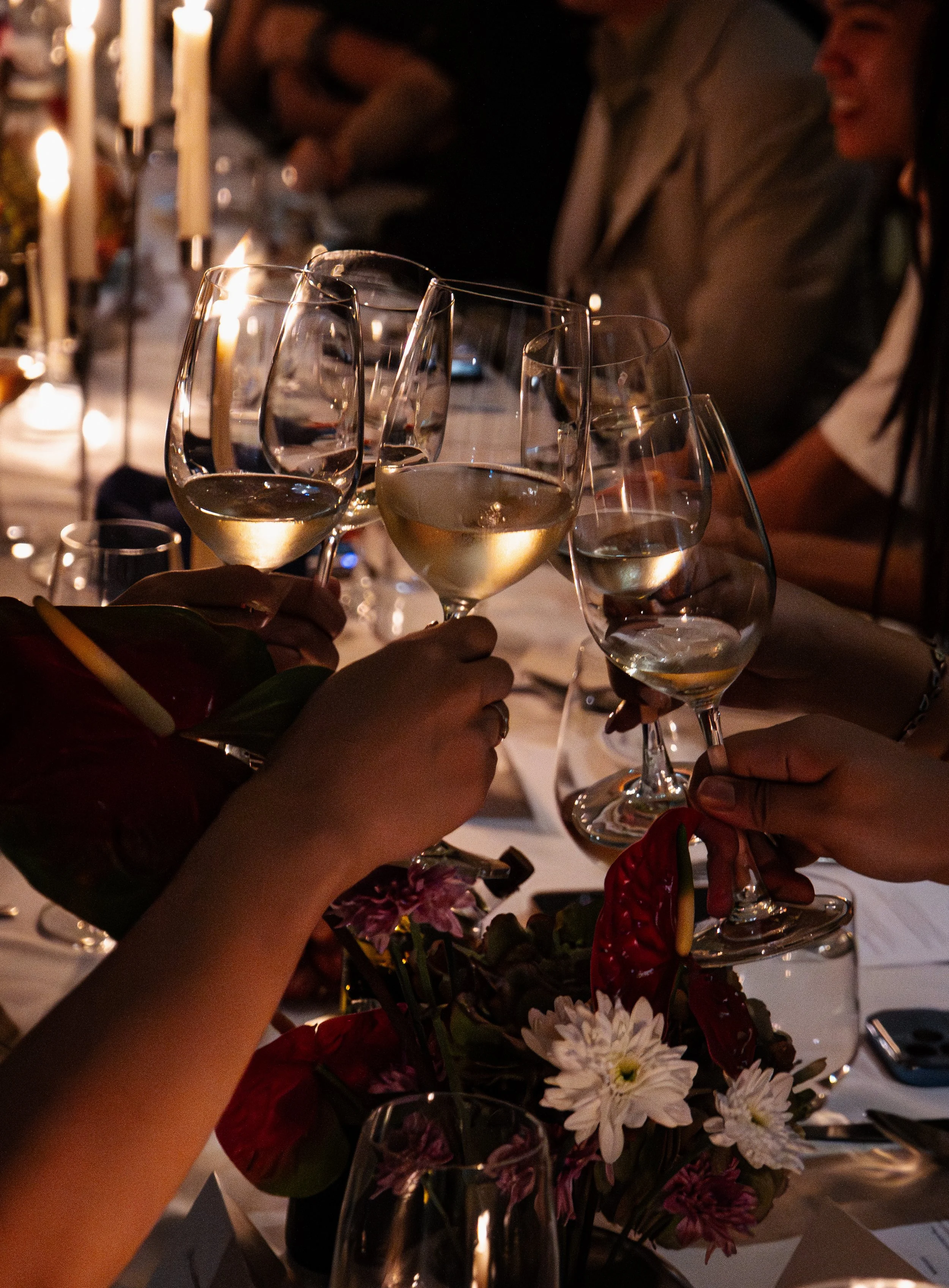 People raising glasses of white wine in a toast at a dinner party, with dimly lit candles and floral decorations on the table.