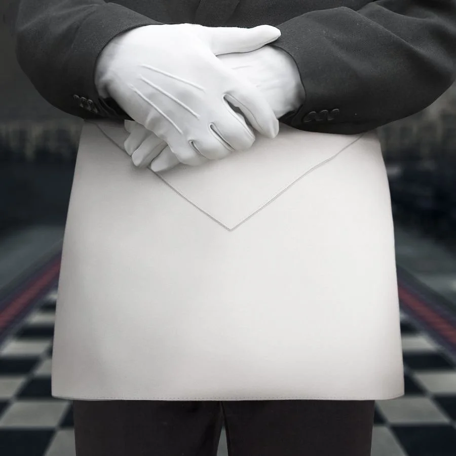 A person wearing a black suit and white gloves, standing on a checkered floor, wearing an apron.