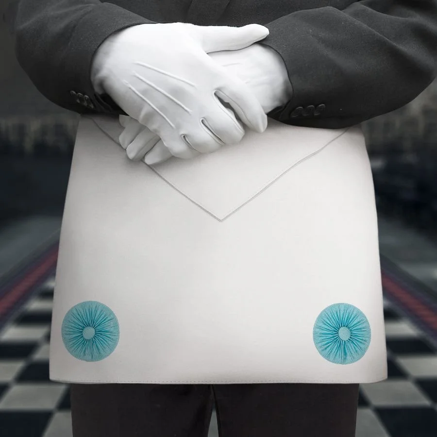 A person wearing a black suit and white gloves, standing on a checkered floor, wearing an apron.