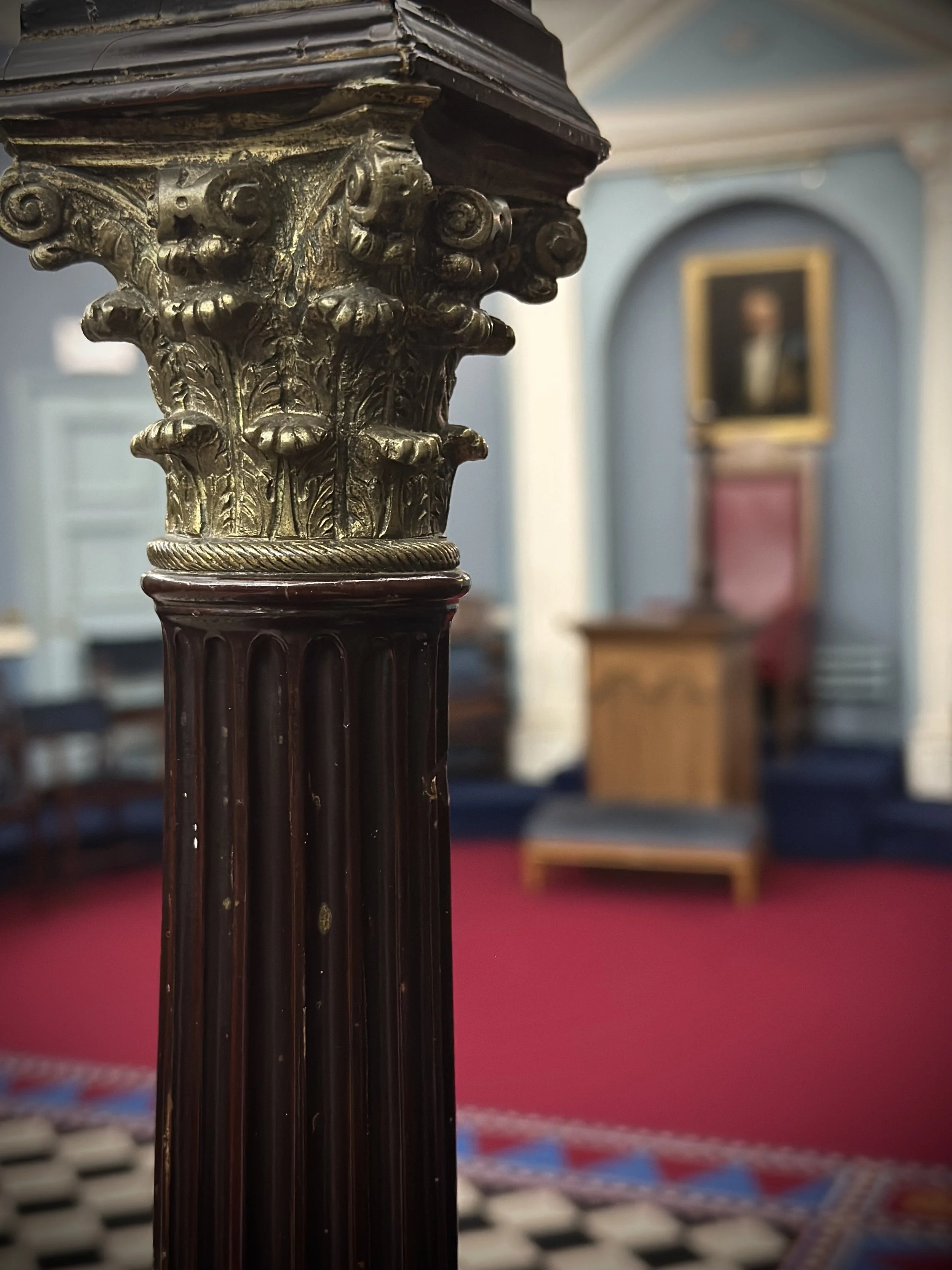 Close-up of an ornate wooden candlestick with gold decorative elements in a room with blue walls, a portrait painting, and historic furniture.