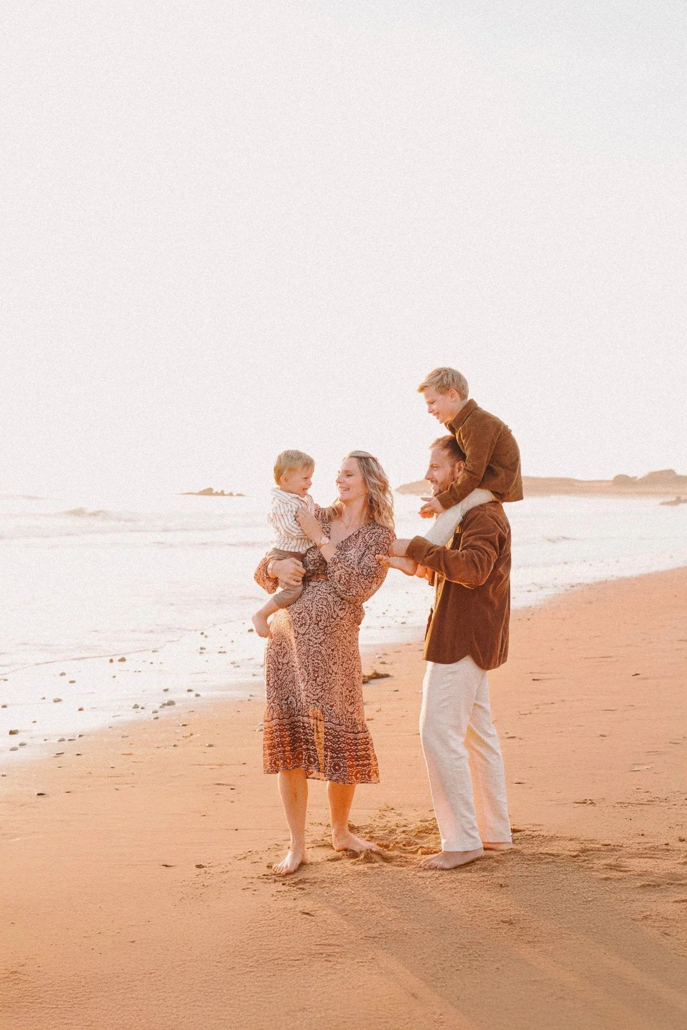 Une famille souriante marche sur la plage au coucher du soleil, avec un père portant un garçon sur ses épaules et une mère tenant la main d'une fille.