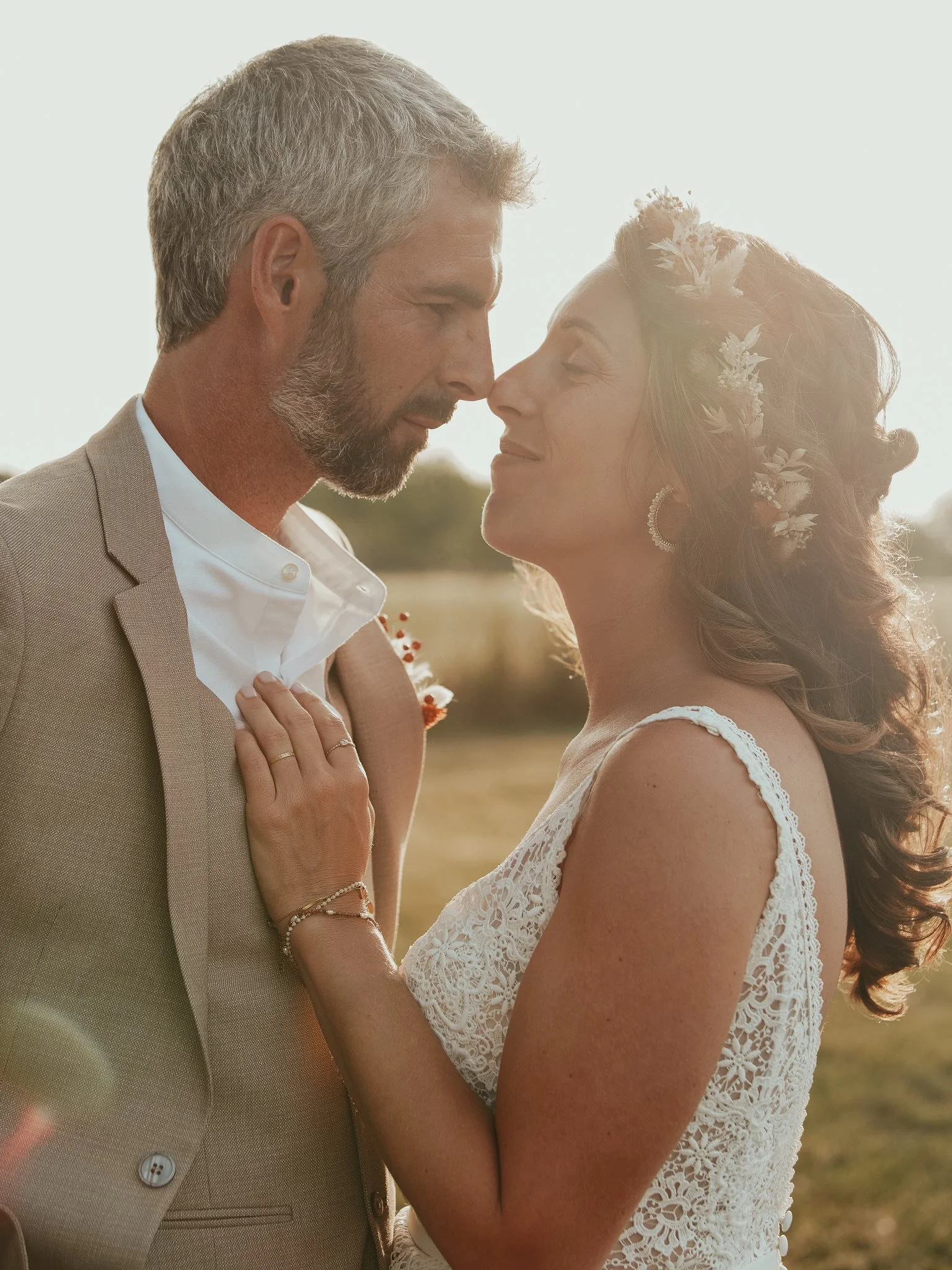 Un couple de mariés s'enlace dans un champ lors d'un coucher de soleil, avec la mariée portant une robe en dentelle et une couronne de fleurs, et le marié portant un costume beige.