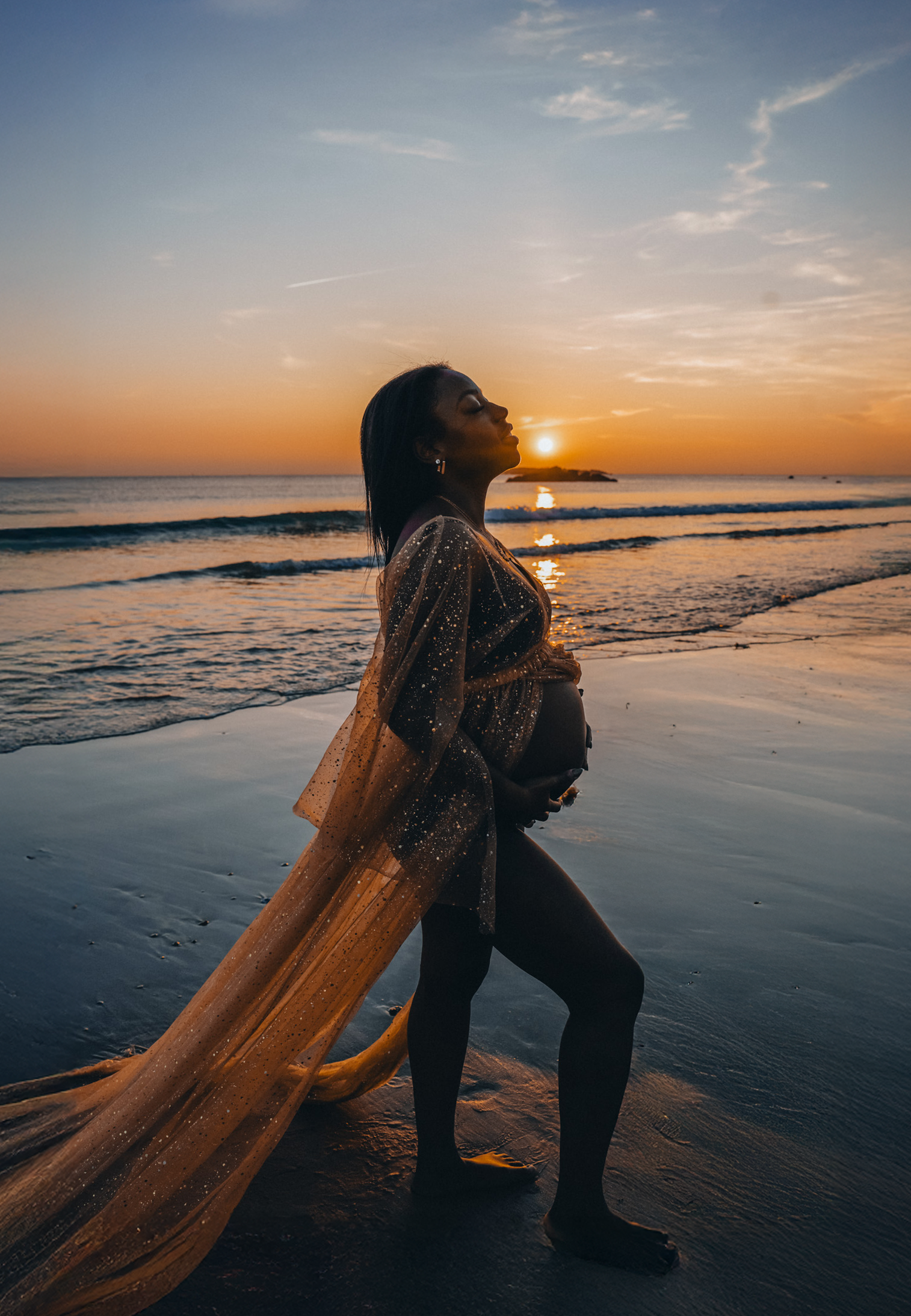 Une femme enceinte se tient sur la plage au coucher du soleil, portant une robe transparente brillante.