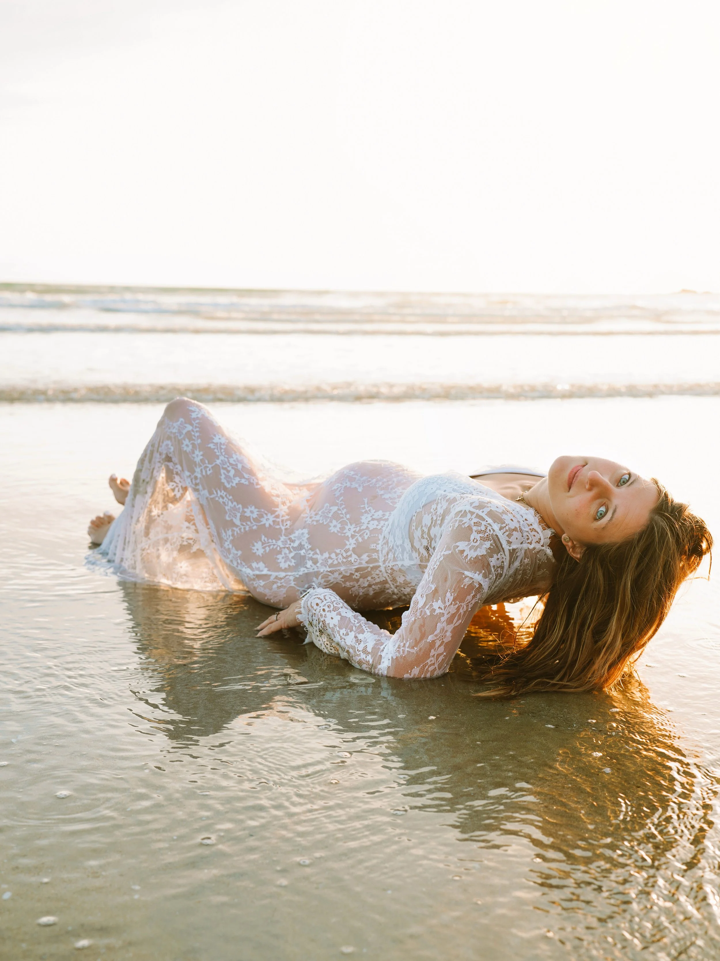 Femme en robe en dentelle blanche allongée sur la plage, face à la mer durant coucher de soleil.