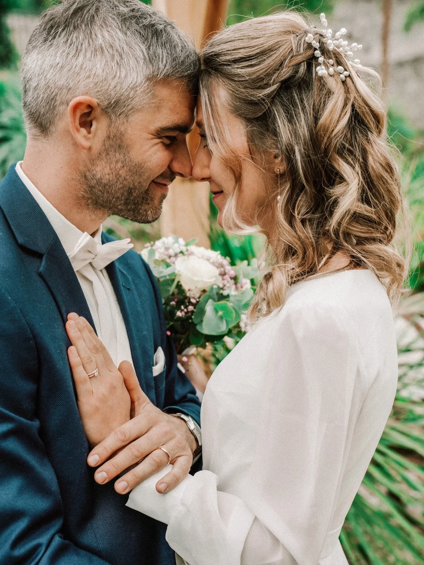 Un couple de mariés s'embrasse tendrement, le front contre front, lors de leur mariage, avec des fleurs en arrière-plan.
