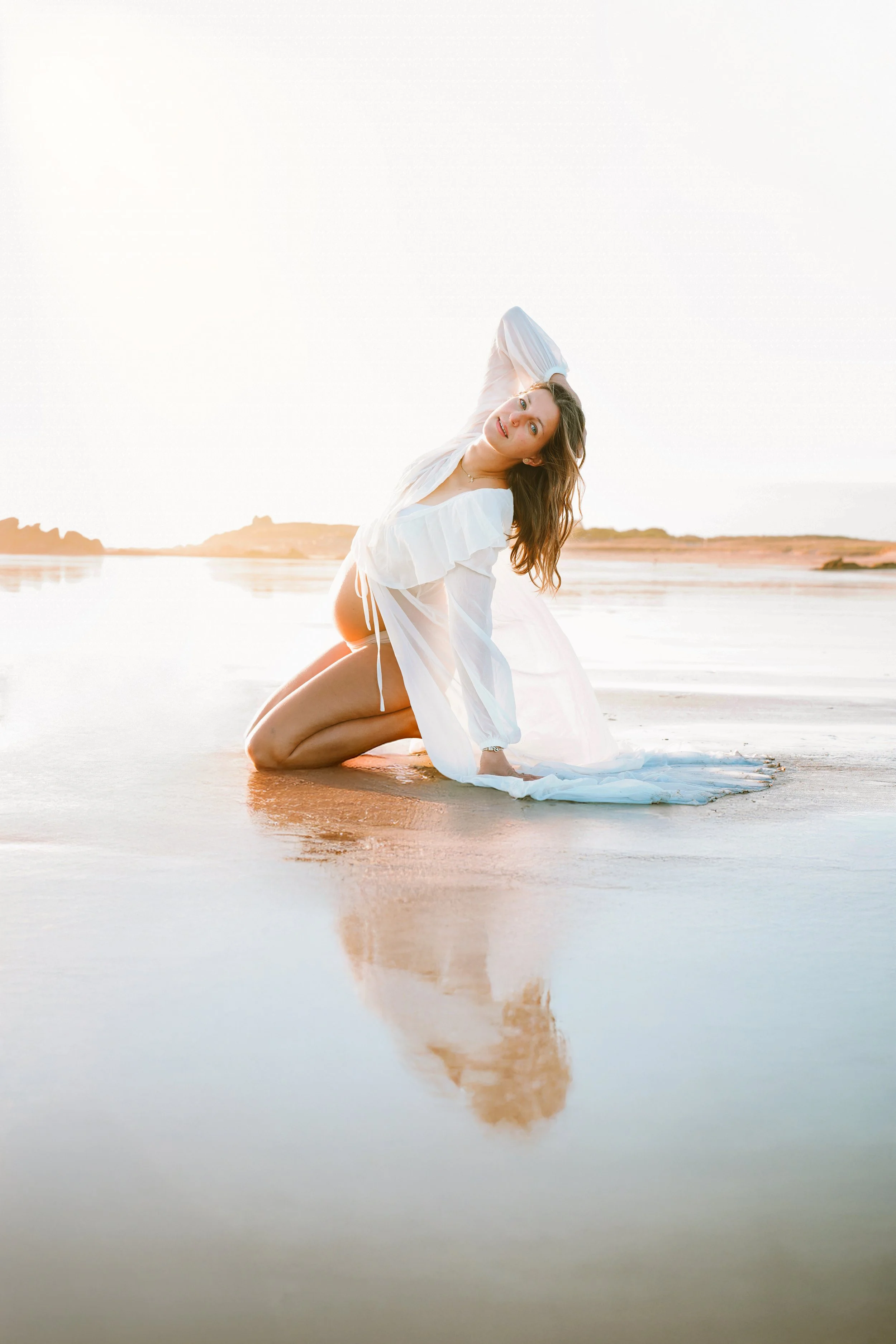Une femme en robe blanche pliée au sol sur la plage, avec reflet dans l'eau, au coucher de soleil.
