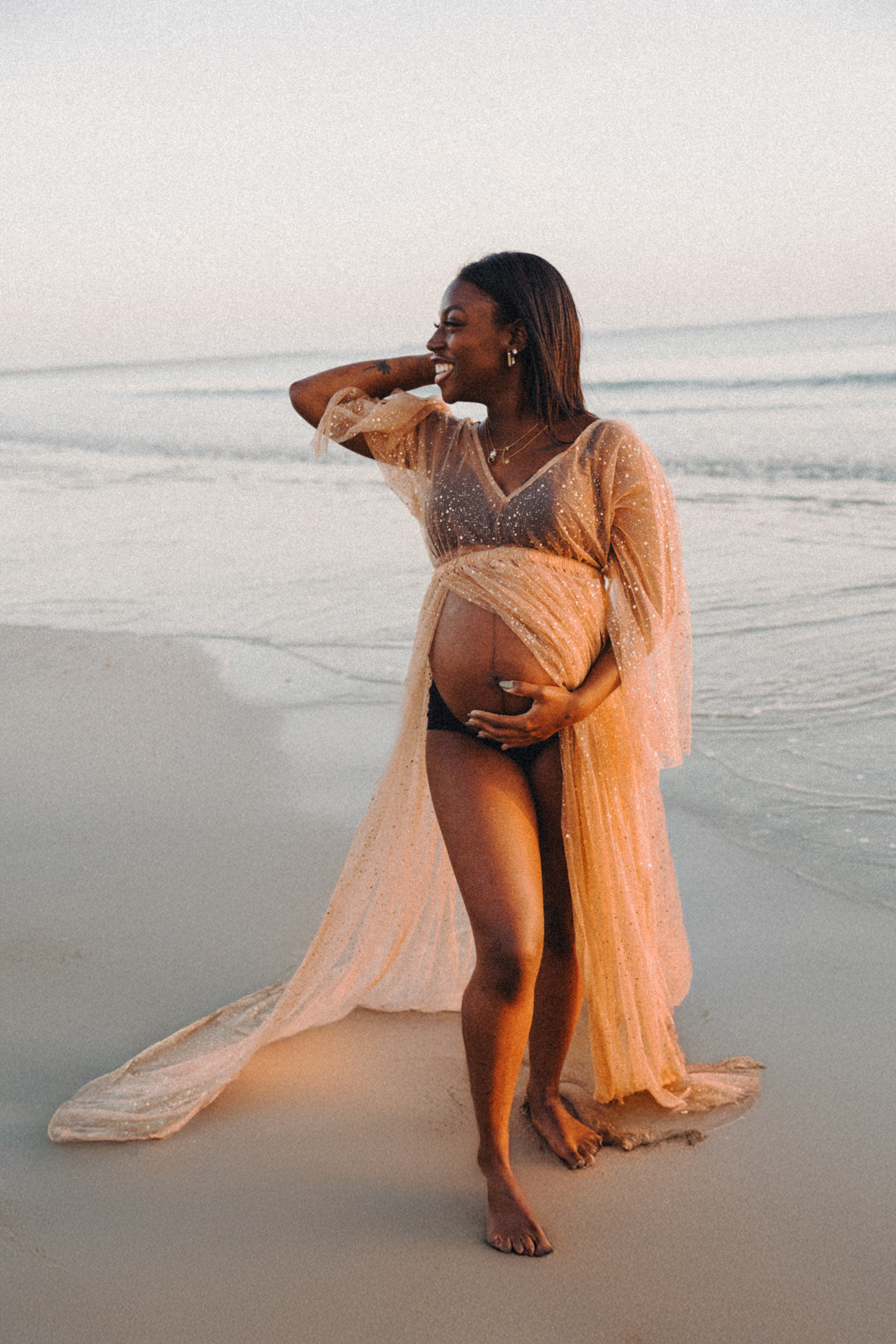 Femme enceinte souriante en robe transparente dorée sur la plage au coucher du soleil