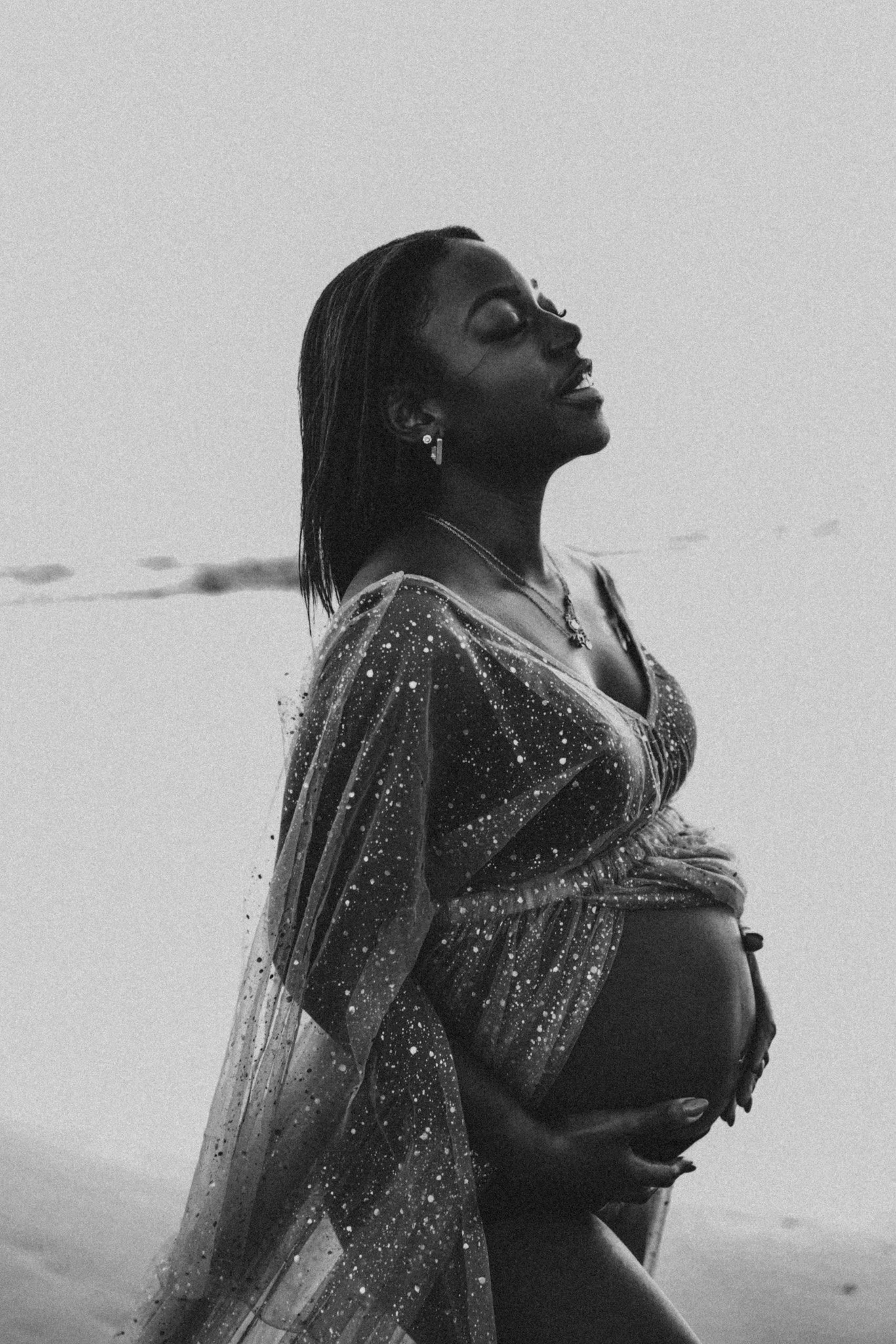 Une femme enceinte en maillot de bain sur la plage, portant une robe en tulle à motifs de galaxie, avec un regard paisible, en noir et blanc.