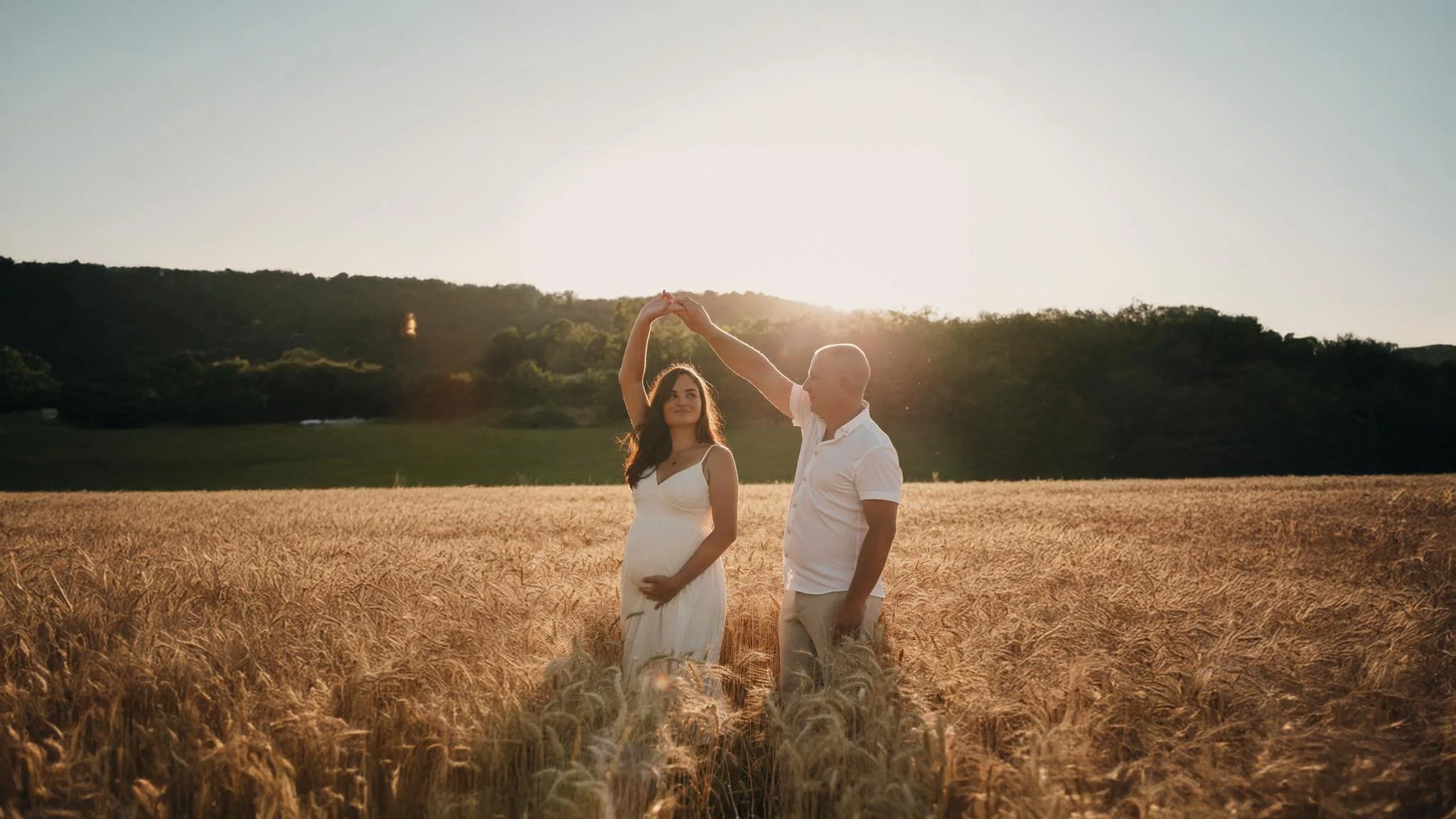 Un couple danse dans un champ de blé doré au coucher du soleil, la femme porte une robe blanche et a la main sur son ventre, l'homme la tient par la main.