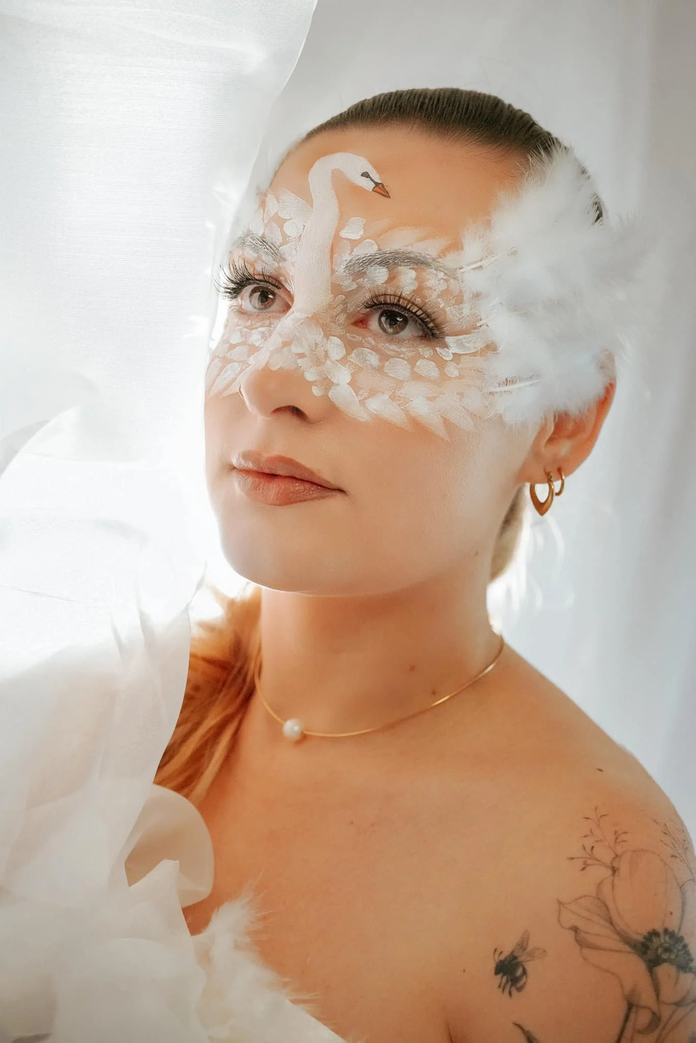 Femme avec un maquillage artistique représentant un cygne blanc, portant des plumes blanches et des bijoux dorés, se tenant devant un rideau blanc.