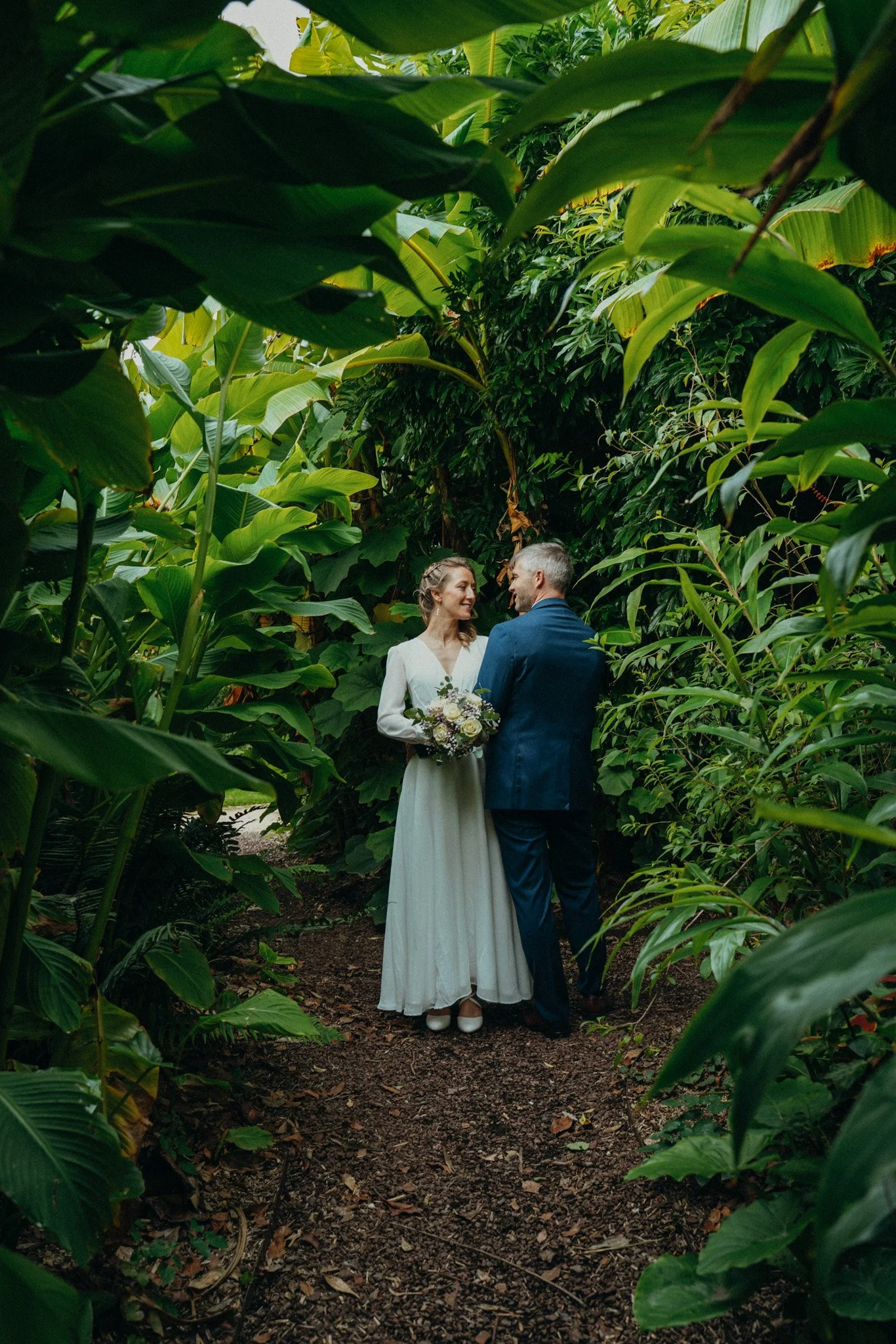 Un couple habillé en mariage debout dans un sentier entouré de végétation dense, elle tient un bouquet, ils se regardent en souriant.