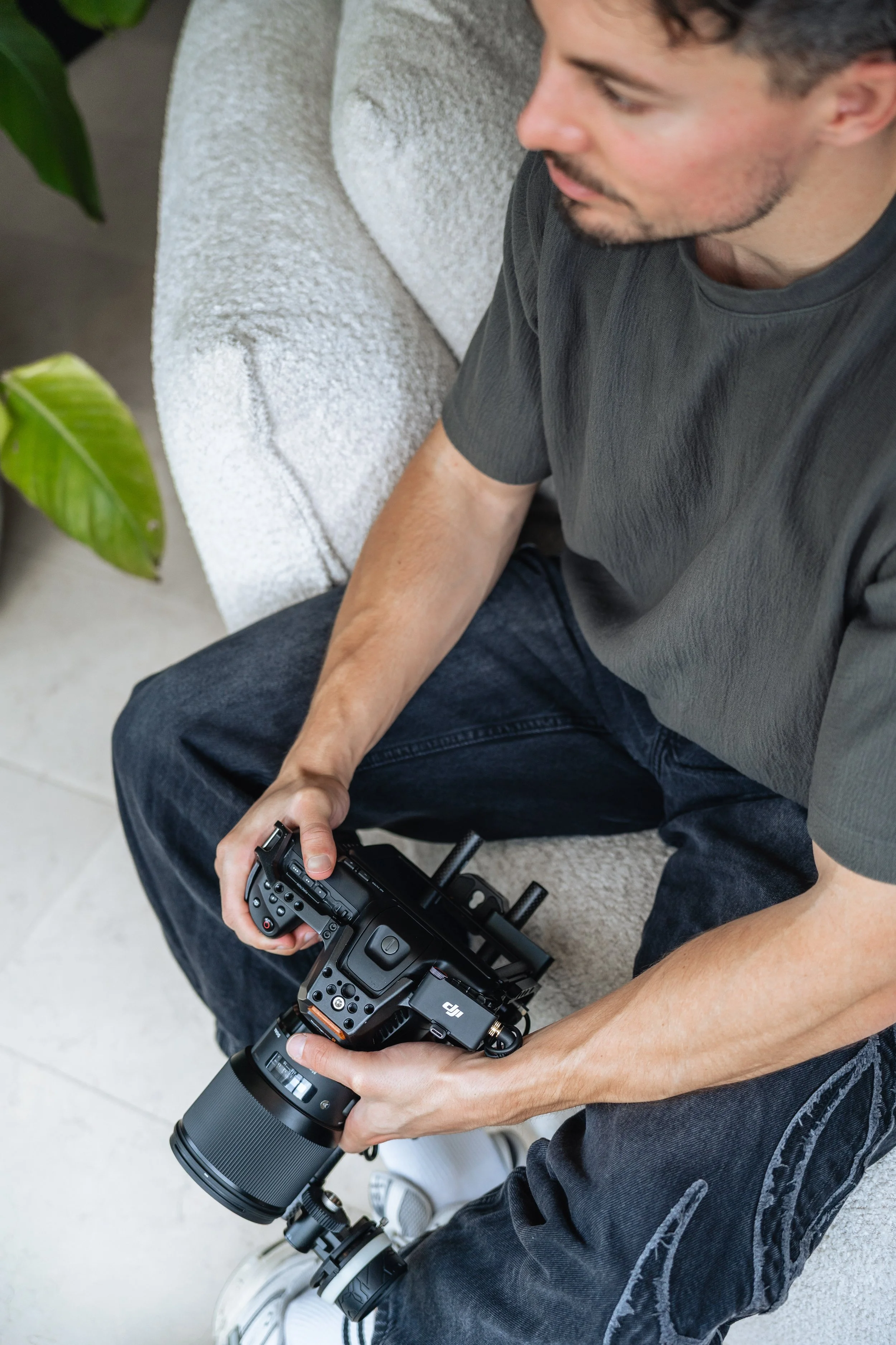 A man sitting outdoors on a light-colored cushioned bench, holding a professional camera with a large zoom lens. He is wearing a dark gray t-shirt and dark jeans, with a leaf of a green plant visible nearby.