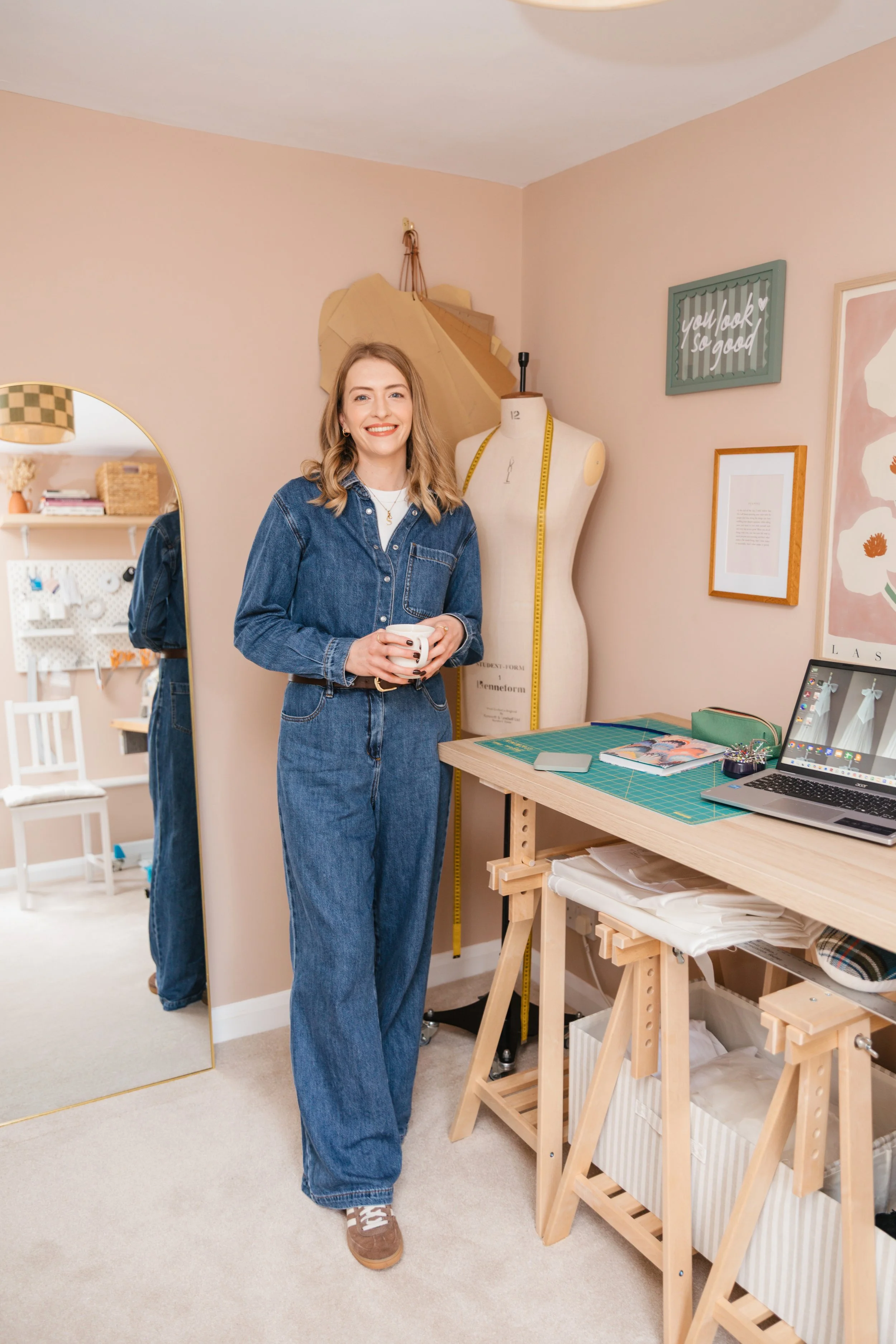 A woman in denim outfit standing in a craft room, holding a mug, smiling, with a dress form, mirror, and sewing materials around her.