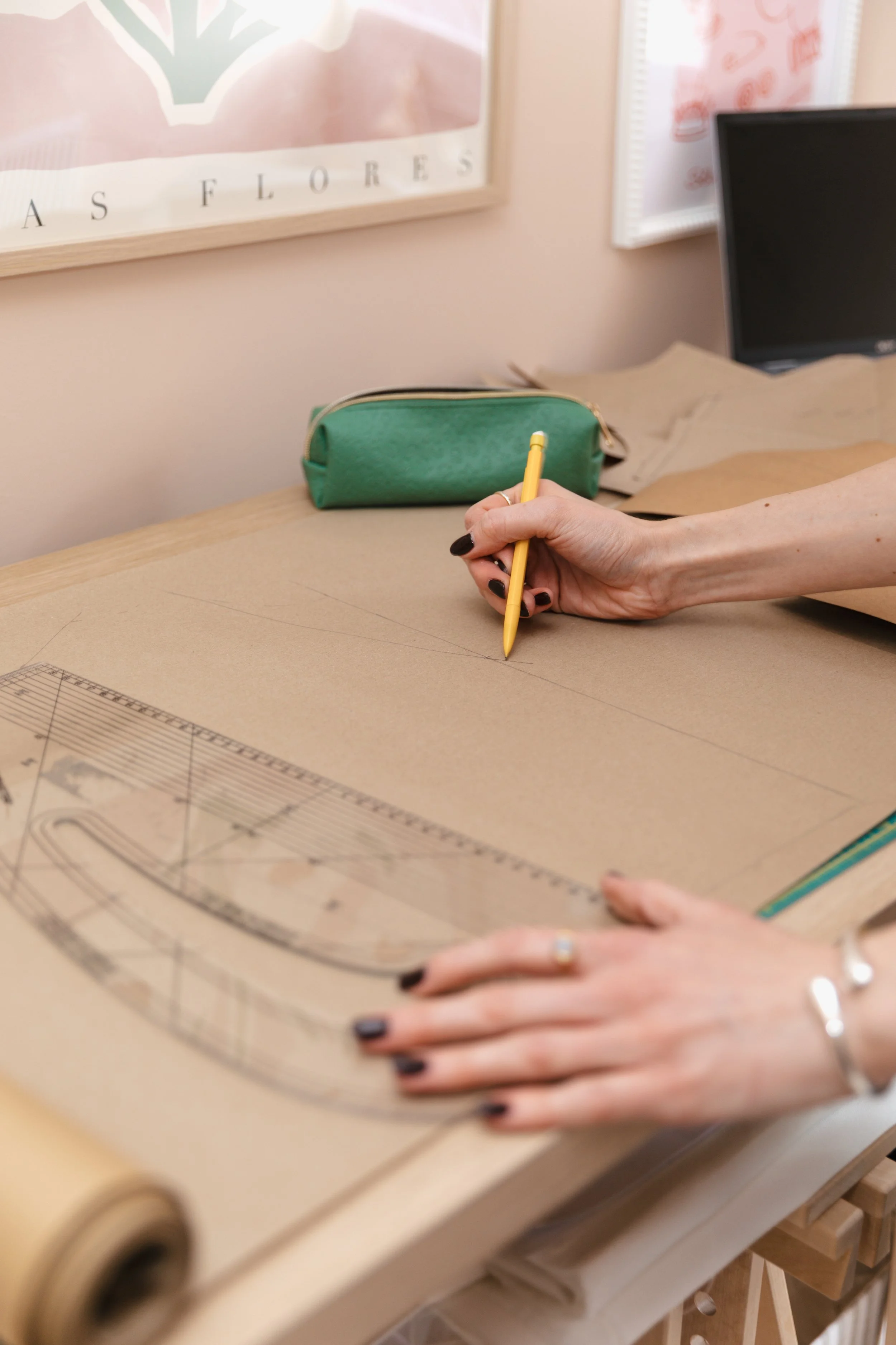 A person drawing with a yellow pencil on a large sheet of brown paper, with a ruler and a protractor nearby, on a wooden table in a workspace.