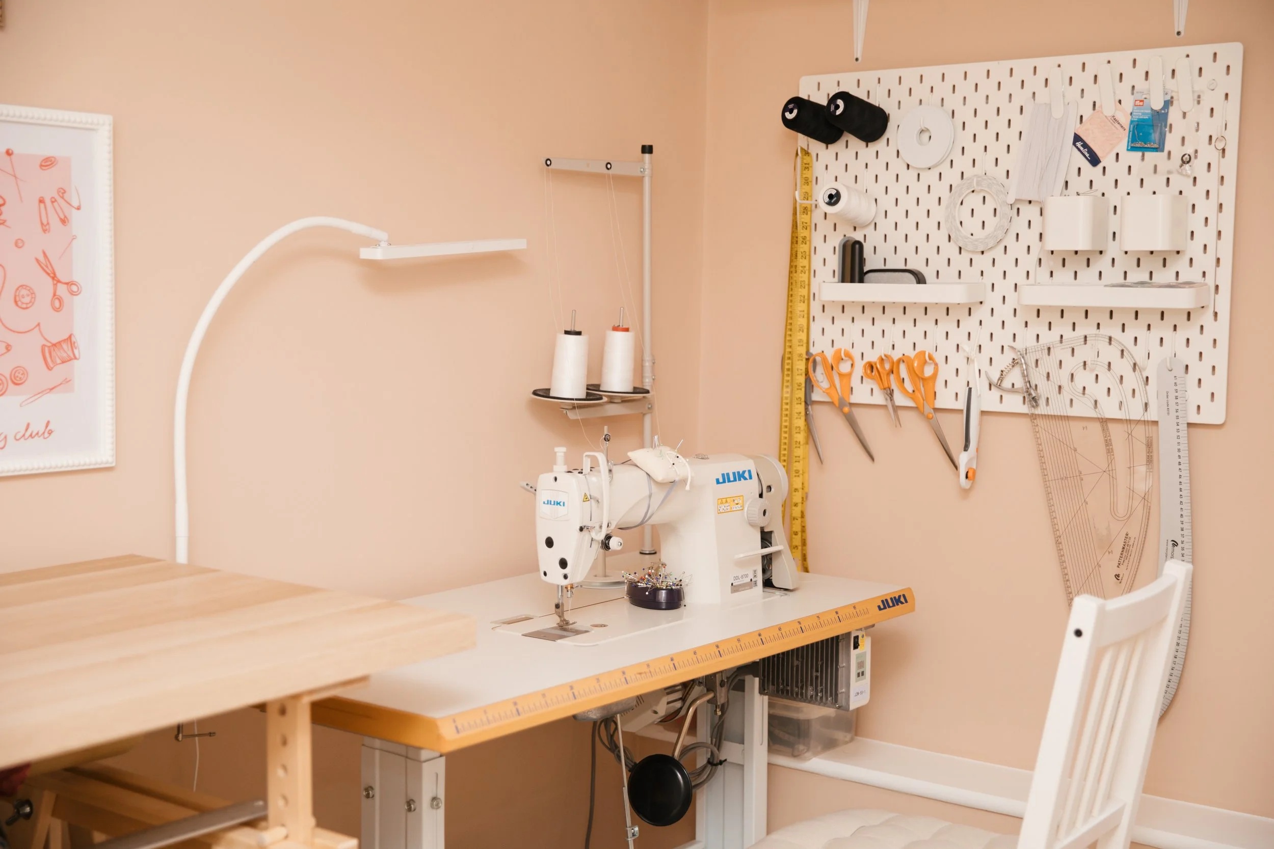 A sewing workspace with a JUKI sewing machine on a worktable, a pegboard with scissors, rulers, and sewing supplies on the wall, and a wooden tabletop in the foreground.