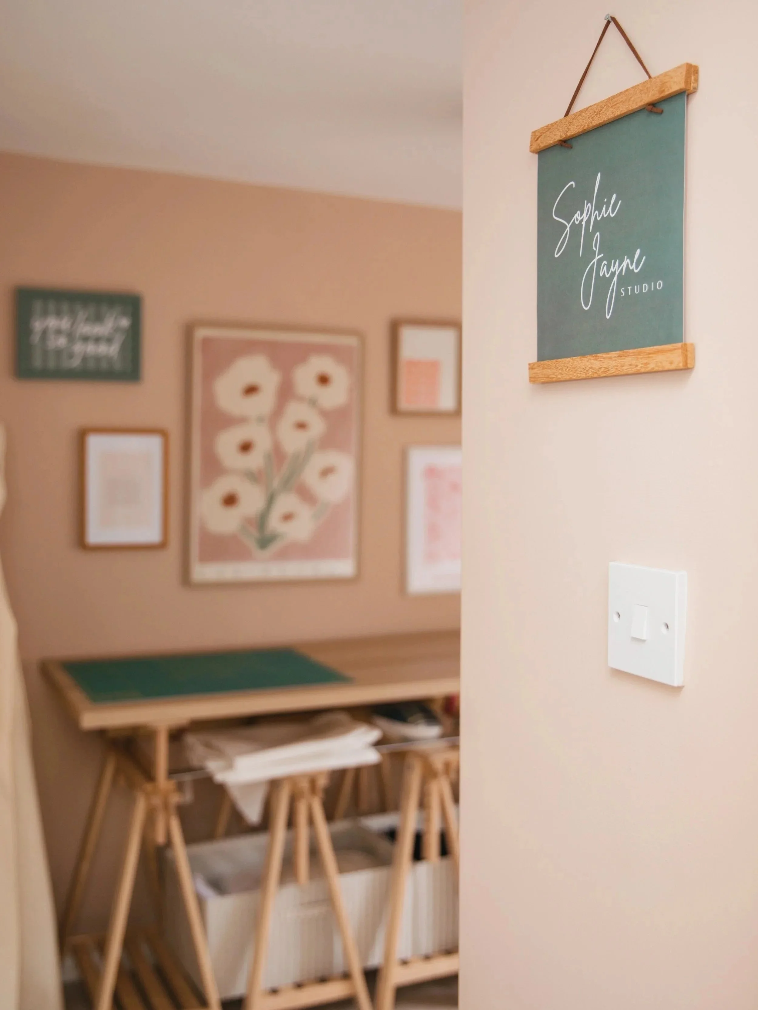 Decorative wall with framed art, a focus on a sign that reads 'Sophie Jayne Studio' hanging on the wall; interior of an art or craft studio with a table and chairs.