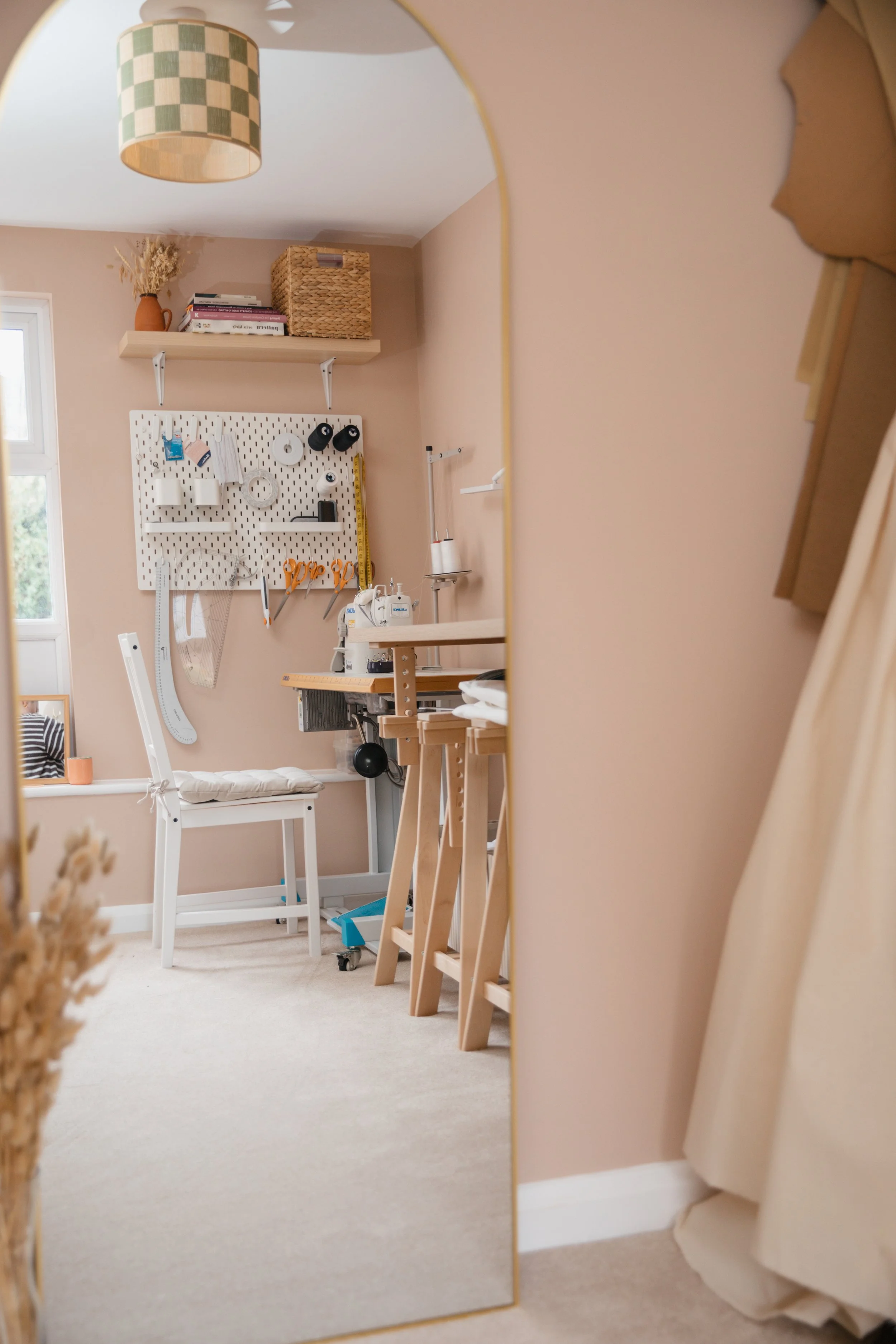 A sewing or craft room reflected in a tall, curved mirror. The room has a pegboard on the wall with various tools and supplies, a white chair, a work table with a sewing machine, and a shelf with storage baskets and books.