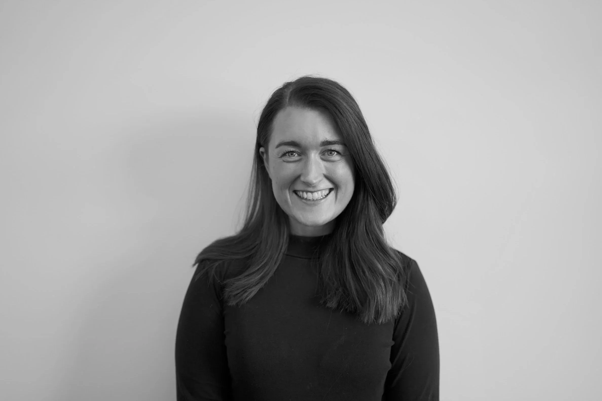 Black and white photo of a smiling woman with long hair wearing a dark top, standing against a plain wall.