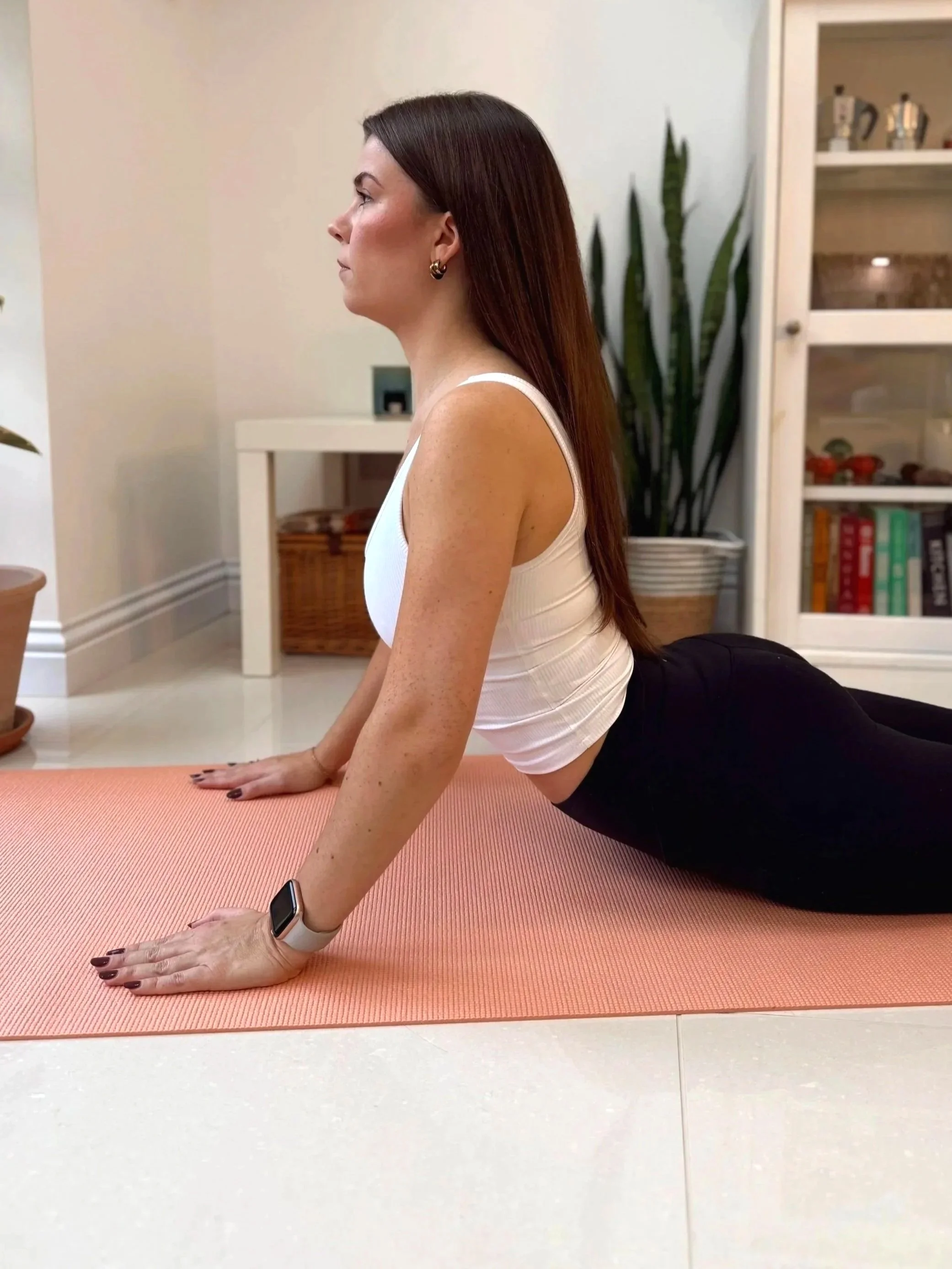 A woman in a white tank top and black pants is practicing yoga or a stretching exercise on a pink yoga mat in a living room.
