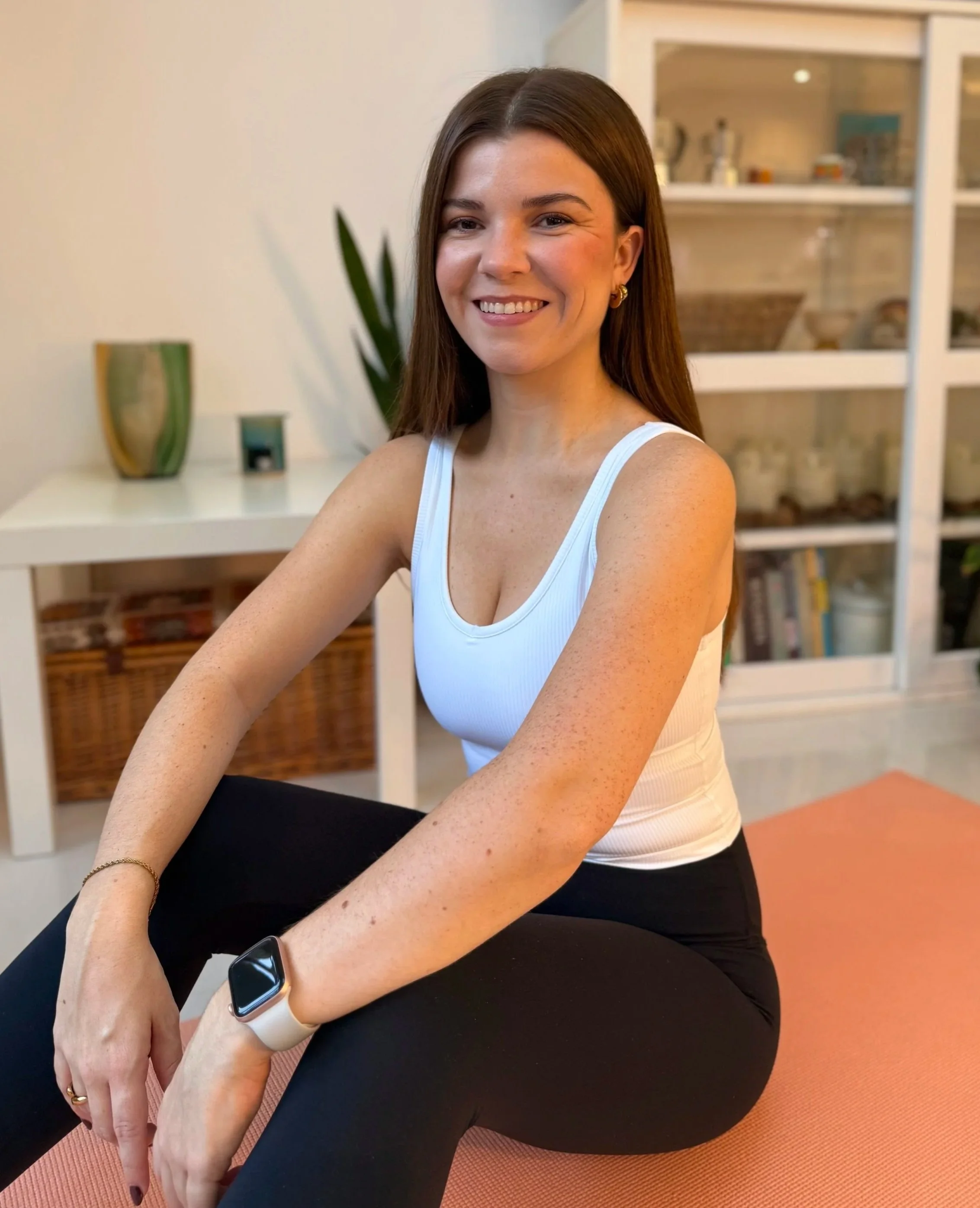 A woman with brown hair, wearing a white tank top and black leggings, sitting on a pink yoga mat in a cozy living room. She is smiling and wearing an Apple Watch on her left wrist.