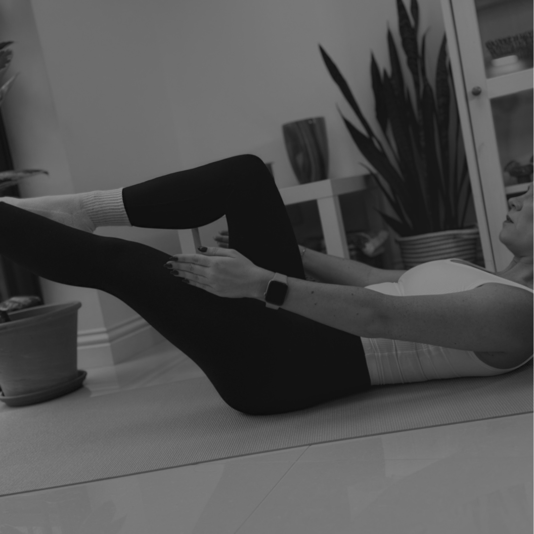 A woman exercising indoors on a yoga mat, lying back with her legs raised and hands on her thighs.