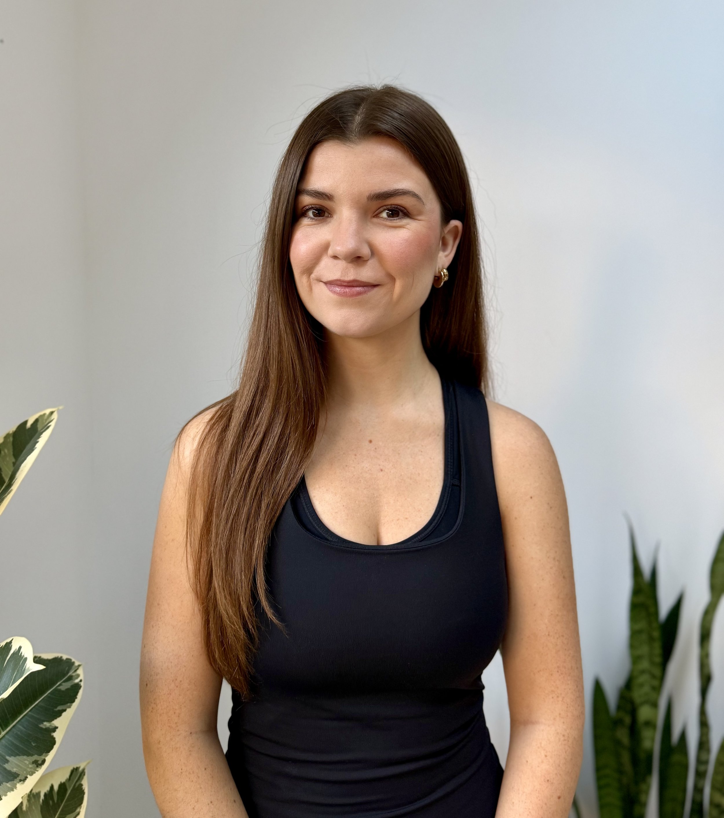 brunette woman in black tank top smiling for camera in front of white background with two green plants in the foreground