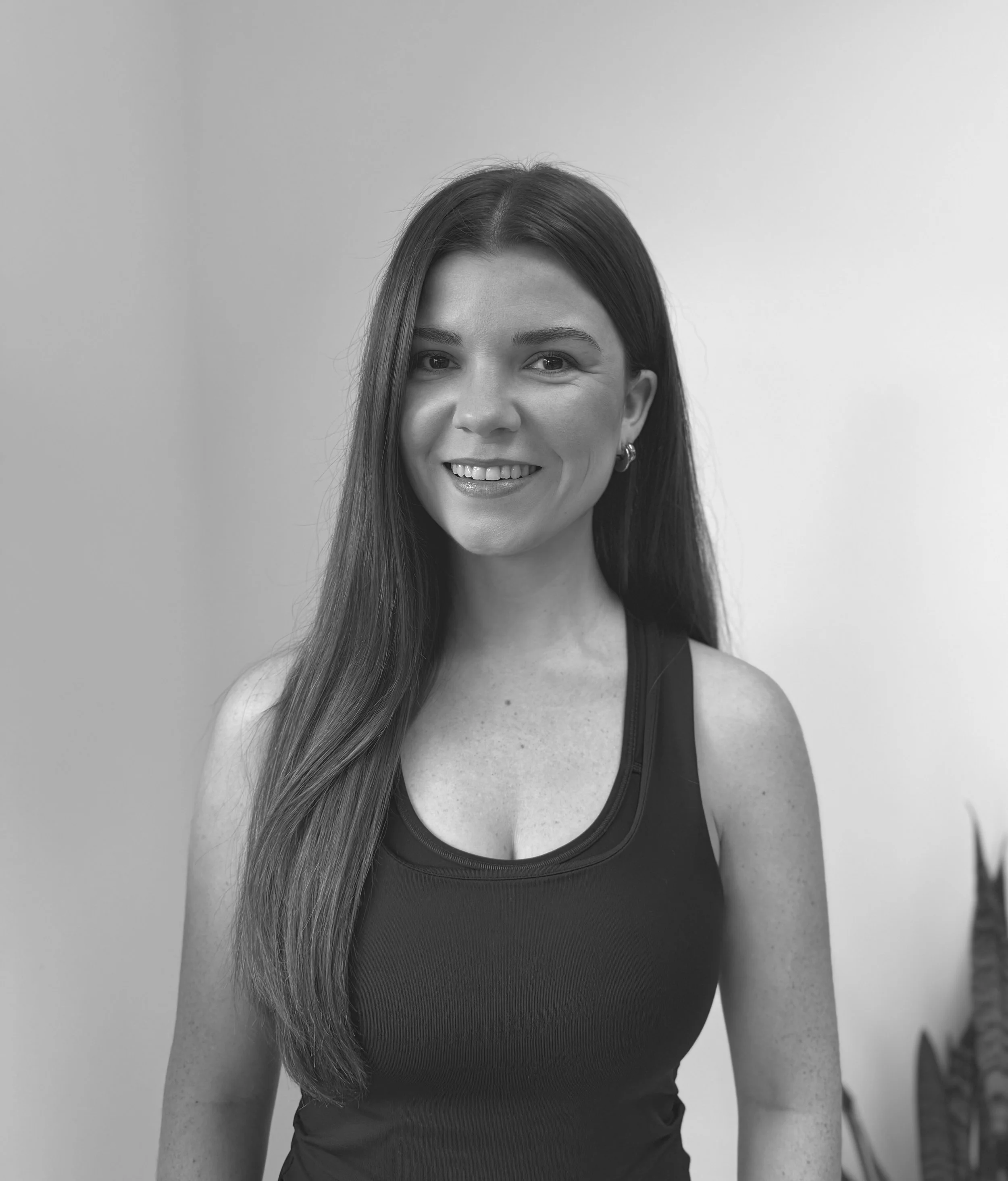 A woman with long dark hair, smiling, wearing a sleeveless top, and standing indoors with a plain background and a potted plant to her right.