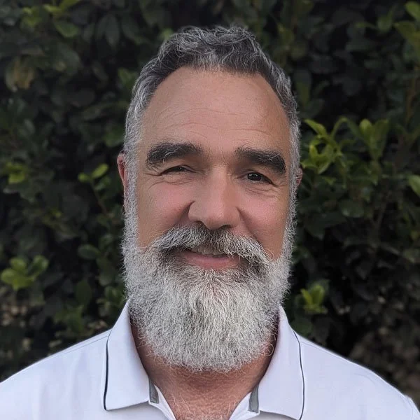 A smiling middle-aged man with a full gray beard and short gray hair, wearing a white collared shirt, standing in front of green leafy bushes.