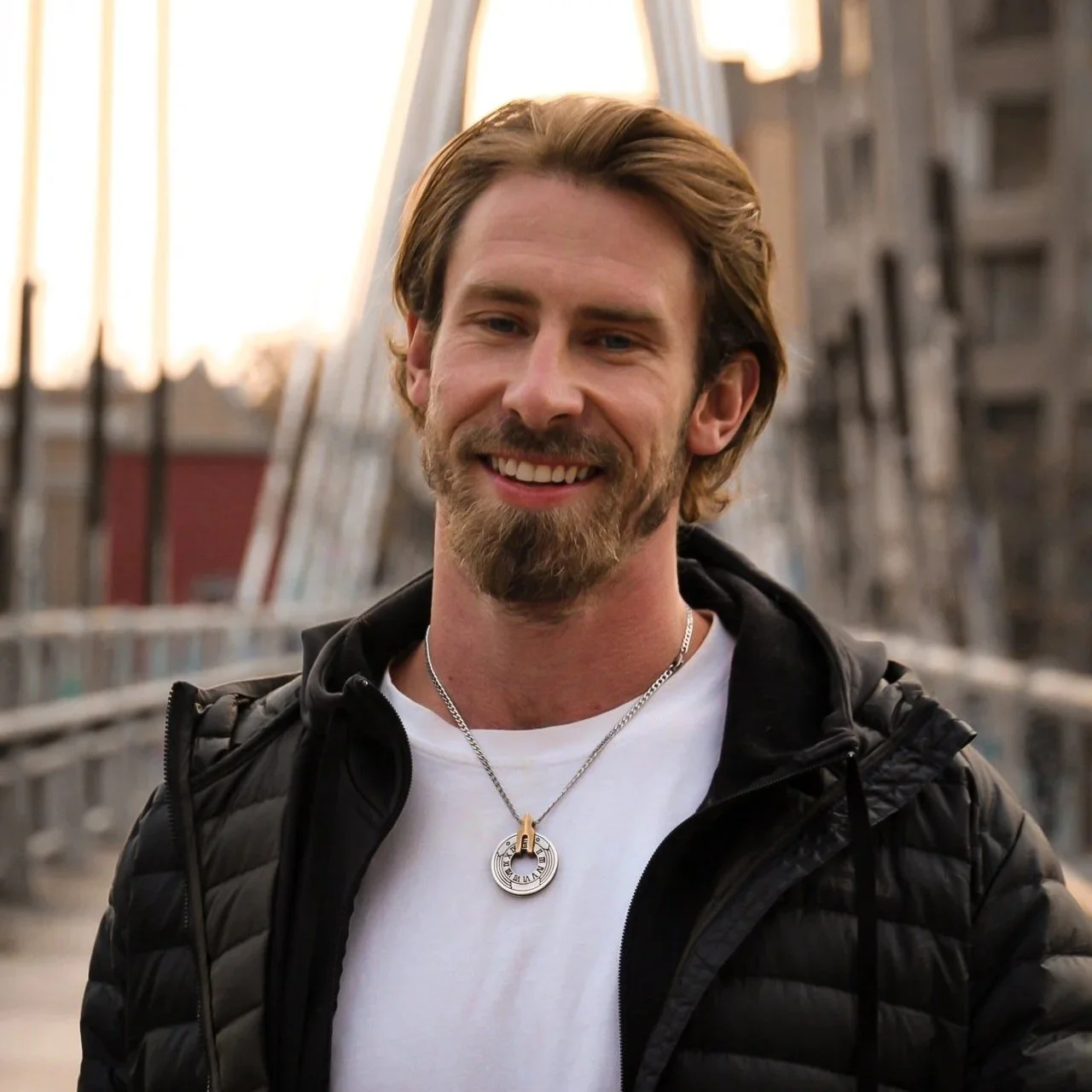 A man with brown hair and a beard smiling outdoors during sunset, wearing a black jacket and a pendant necklace.