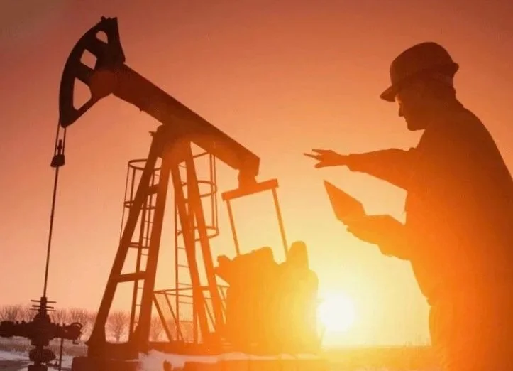 Silhouette of a man in a hard hat holding a tablet, standing near an oil pumpjack during sunset.
