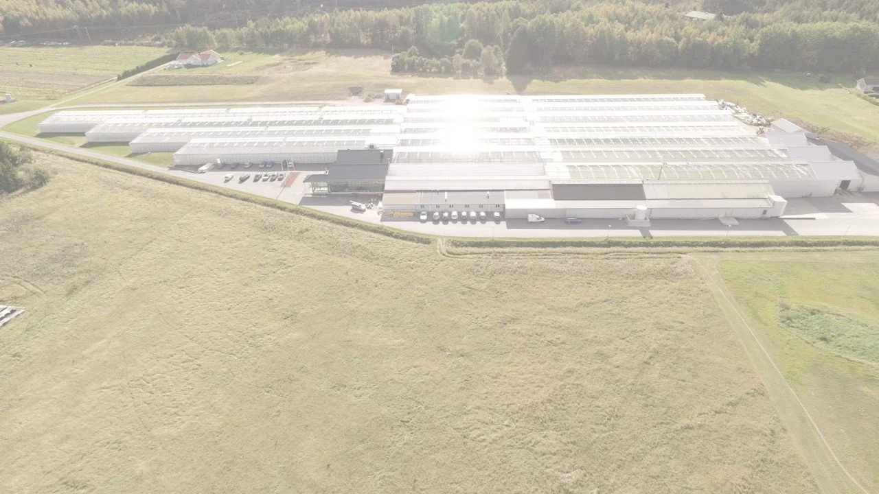 Flygbild av ett modernt, stort växthus med flera byggnader, parkeringsplatser och omgivande gröna fält och skog.