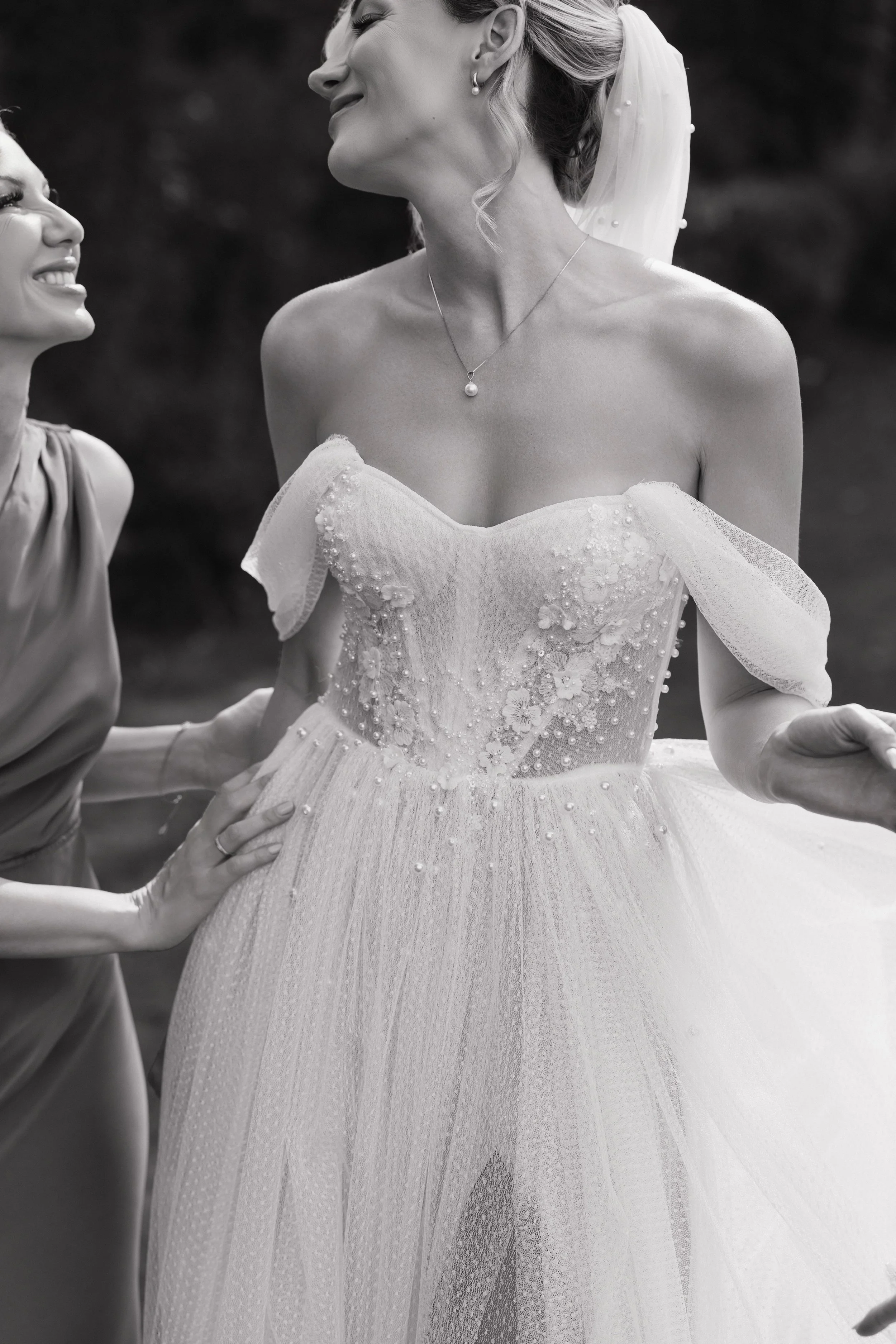 Black and white photo of a bride in a wedding dress smiling and holding hands with a woman, likely a bridesmaid, outdoors.