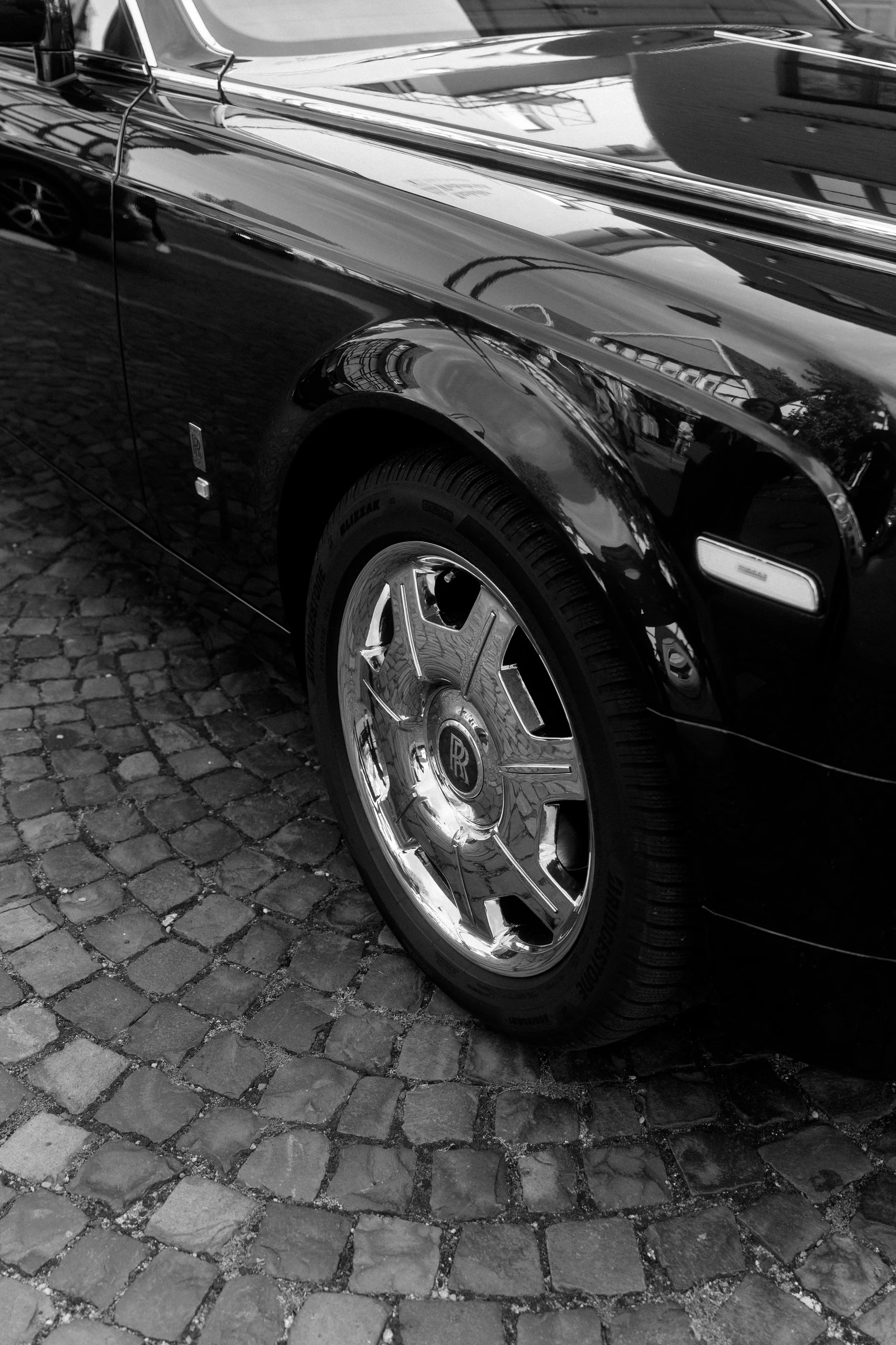 Close-up of a black vintage Rolls-Royce car with a shiny chrome wheel, parked on a cobblestone street, reflected in its polished surface.