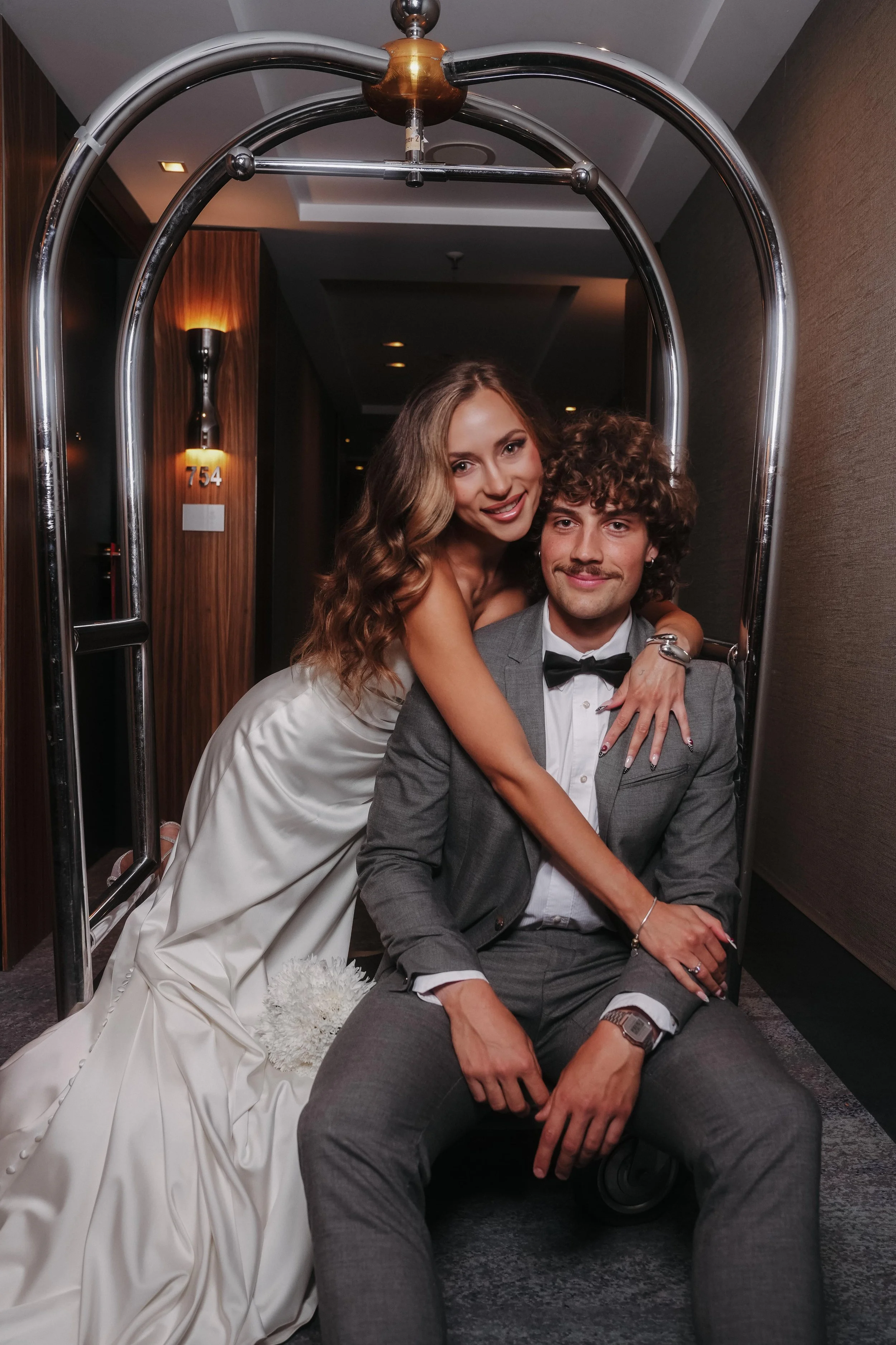 A couple dressed in formal attire, the woman in a white wedding gown, poses inside a luggage cart, smiling and embracing each other in a hotel hallway.